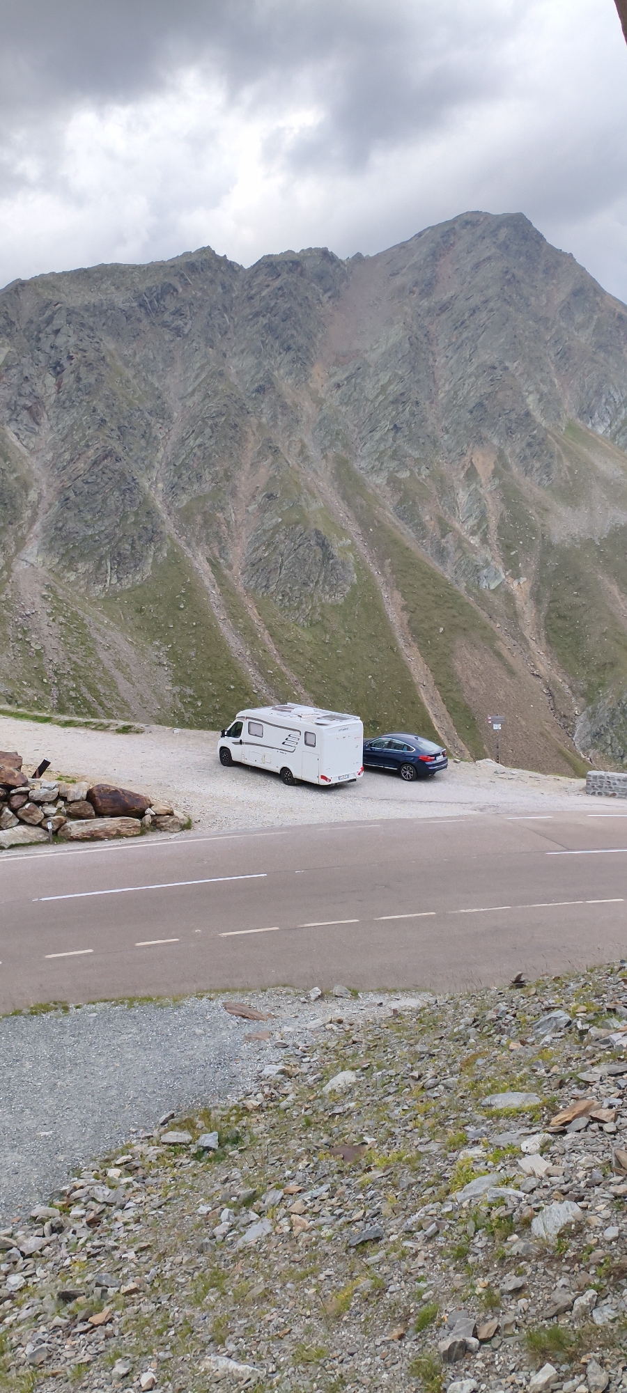 The image shows a panoramic view of a mountainous landscape with a cloudy sky overhead. In the foreground, a paved road with white lane markings cuts across the scene. To the left of the road, a rocky embankment is visible. In the middle ground, parked on a gravel shoulder adjacent to the road, is a white camper van and a dark-colored sedan. The camper van is facing to the right, and the sedan is parked behind it, facing slightly to the left. Further back, the steep, rocky slopes of a mountain rise dramatically, with patches of green vegetation sparsely dotting the terrain. The overall environment suggests a high-altitude, remote location, likely a mountain pass or scenic viewpoint. The location is identified as being in the vicinity of Sölden, Austria, a popular ski resort town known for its alpine scenery. There are no people visible in the image.