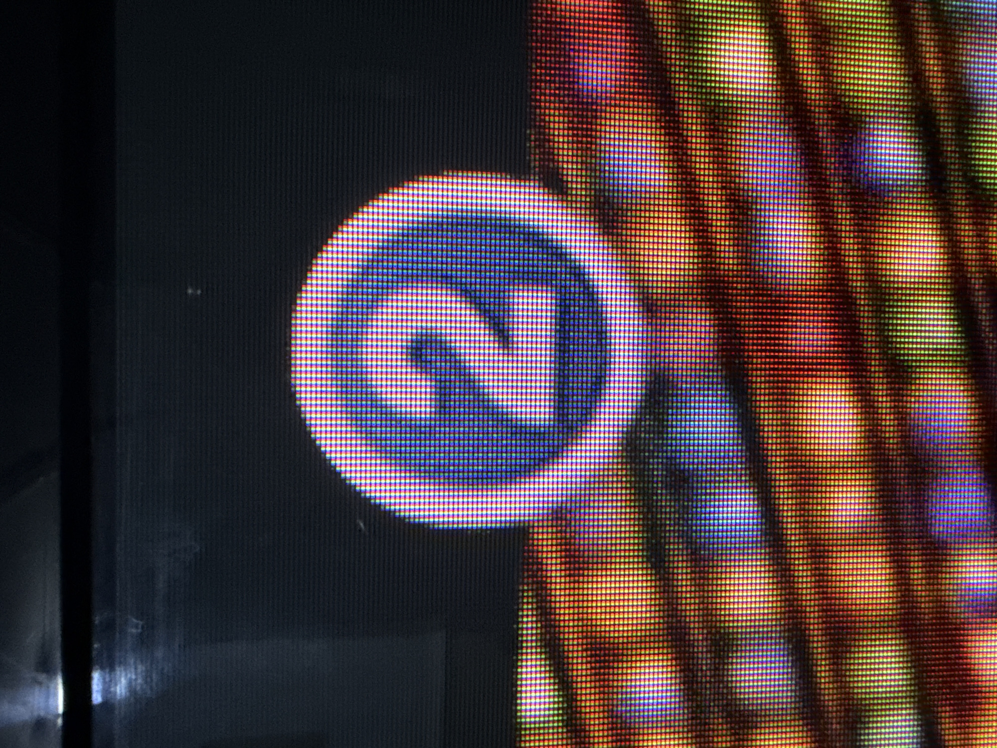 The image shows a close-up of a digital display, likely a television screen or monitor, exhibiting a pixilated representation of the number "2" within a blue circular emblem. The emblem is centered on the upper portion of the screen, against a dark background. Below the emblem, the screen displays a blurred, colorful pattern of lights and shapes in shades of orange, red, yellow, and blue, suggesting a dynamic or abstract visual element. The extreme close-up reveals the individual red, green, and blue sub-pixels that constitute the image on the display. There are no discernible people, actions, or environmental details outside of the digital display itself. The location is unknown.