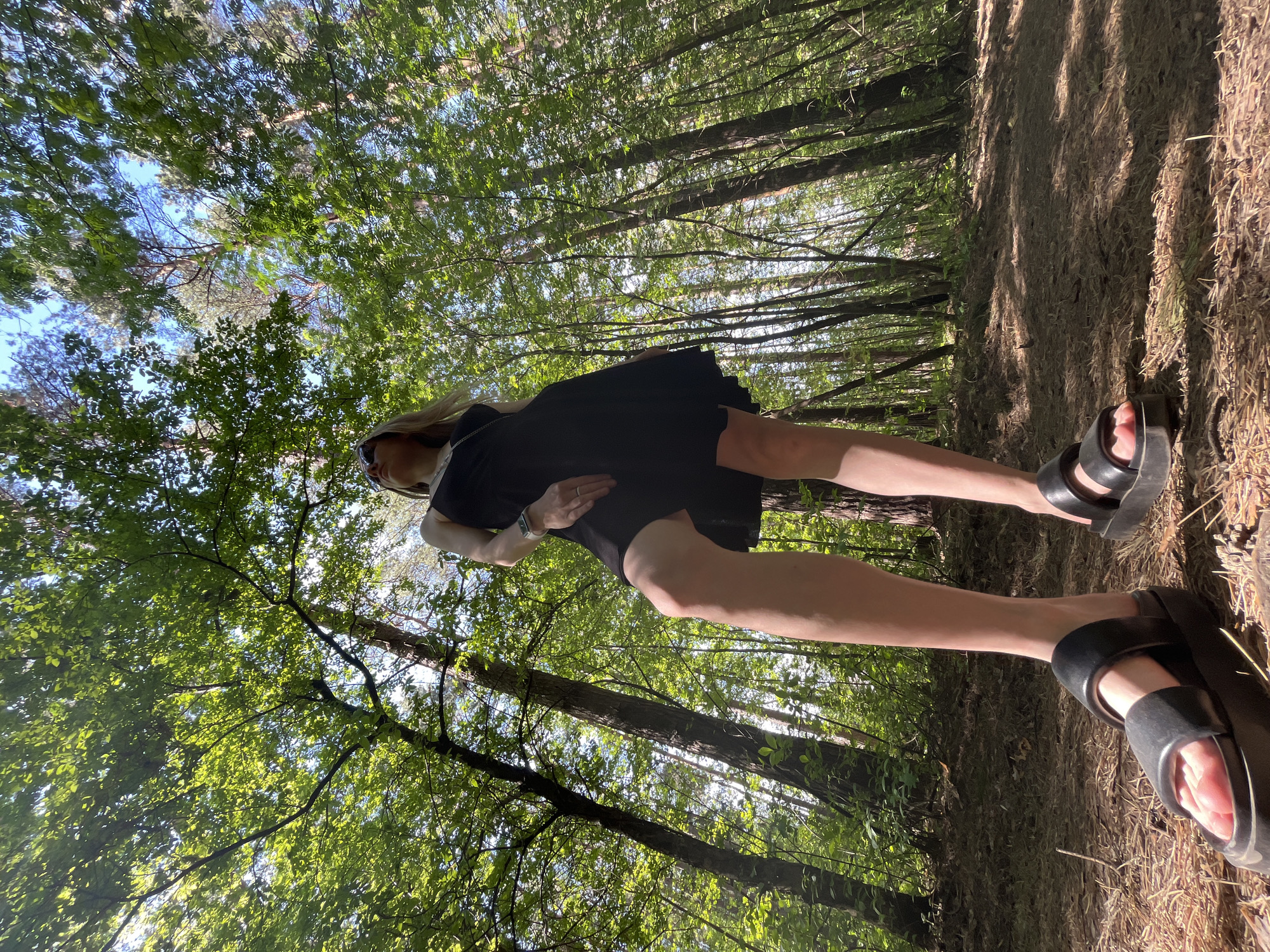 The image shows a person standing on a path in a forest. The person is wearing a black dress and black sandals. Trees surround the path and the sky is visible through the canopy. The ground is covered with leaves and dirt. The picture was taken at a low angle, looking up at the person. The location is within the city limits of Chaikovsky, Russia.