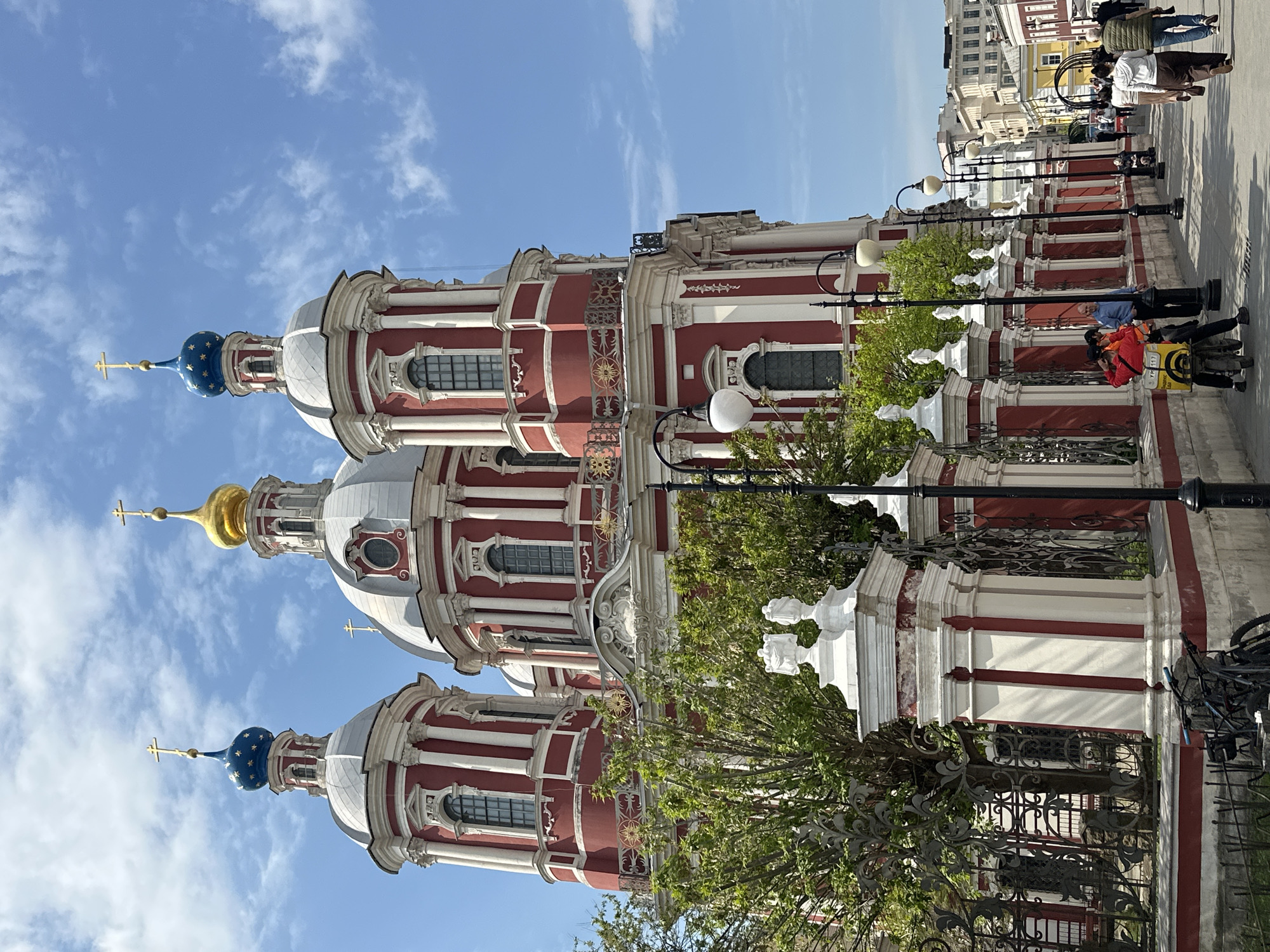 The image depicts the Church of St. Nicholas in the Pyzhi in Moscow, Russia. It is a baroque-style church with red and white exterior. The church has three prominent domes: two are blue with gold stars, and the central one is gold. The sky is light blue with scattered clouds. In the foreground, there is a fence made of black wrought iron. There are also trees with green leaves partially obscuring the view of the church. On the right side of the image, a sidewalk with pedestrians is visible. One individual in a red jacket and yellow backpack appears to be on a scooter. There are also lampposts that run along the sidewalk. The architectural style and colors of the buildings in the background suggest the location is in the historical part of the city.