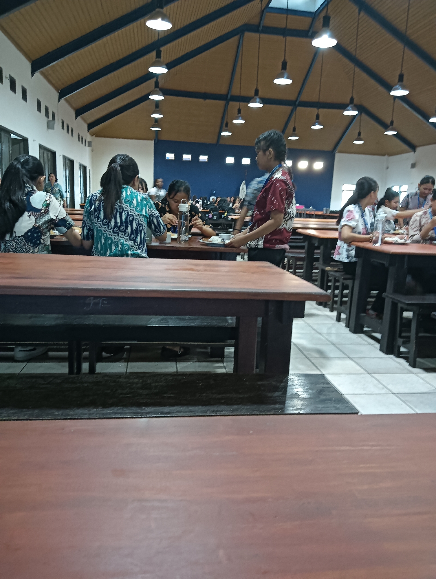 The image shows an indoor scene, possibly a canteen or dining hall, with multiple tables and chairs. Several people are seated at the tables, some eating and others conversing. The architecture features a high, sloped ceiling with wooden panels and exposed beams, illuminated by pendant lights. The walls are a light color, with a dark blue section visible in the background. The floor is tiled with a pattern of light and dark squares.
In the foreground, a long, dark wooden table is prominent. Behind it, other tables are arranged in rows. Several individuals are visible at these tables. On the left side, a woman in a patterned blue and white top is seated. To her right, another woman in a similar patterned blue and green top is also seated. Further to the right, a young man in a red and white patterned shirt is standing, seemingly interacting with those seated at a table. Several other people are visible in the background, some of whom appear to be students. The overall impression is a communal dining area during a mealtime.
The image's context within the city of Tomohon, Indonesia, is not directly discernible from the visual information. However, the clothing and general setting suggest a tropical or sub-tropical location. Without additional information or identifying landmarks, pinpointing the specific location within Tomohon is impossible.