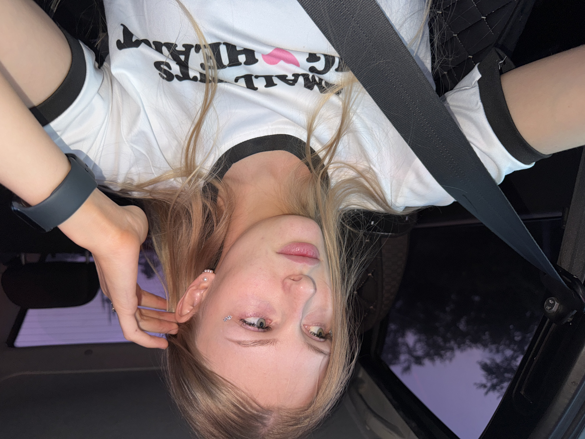 A young woman with blonde hair is sitting in the passenger seat of a car at dusk. She is wearing a white t-shirt with black trim and a graphic print that includes the words "SMALL" and "BIG HEART" along with a pink heart. She has a seatbelt fastened across her chest. Her left hand is touching her hair near her ear, and she is looking out the side window. The window shows a purple twilight sky and silhouetted trees. She is wearing earrings and has some subtle eye makeup, including small rhinestones under her left eye. A dark watch is visible on her right wrist. The interior of the car is dark, with the seatbelt buckle and part of the car seat visible. The image appears to have been taken in the city of Perm, Russia.