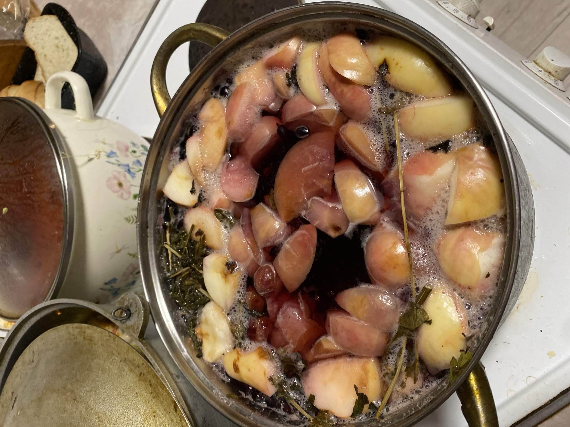 In a kitchen environment, possibly in the city of Chornomorsk, Ukraine, a metal pot filled with a bubbling, dark red liquid is the central focus. The pot contains numerous chopped pieces of what appear to be peaches or apples, some with reddish skins. Dark berries, possibly blueberries or cherries, are also visible throughout the mixture. Sprigs of green herbs are interspersed among the fruit. The pot is situated on a white stovetop, with a knob for temperature control visible in the lower right corner. To the left of the pot, a large, round, metallic lid with a slightly browned surface is partially visible, suggesting another cooking vessel. Behind the pot, another cooking pot with a cream-colored exterior adorned with a floral pattern is present, covered by a dark red lid. In the background, a black container holds slices of bread. The overall scene depicts food preparation, likely the cooking of compote or a similar fruit-based beverage.