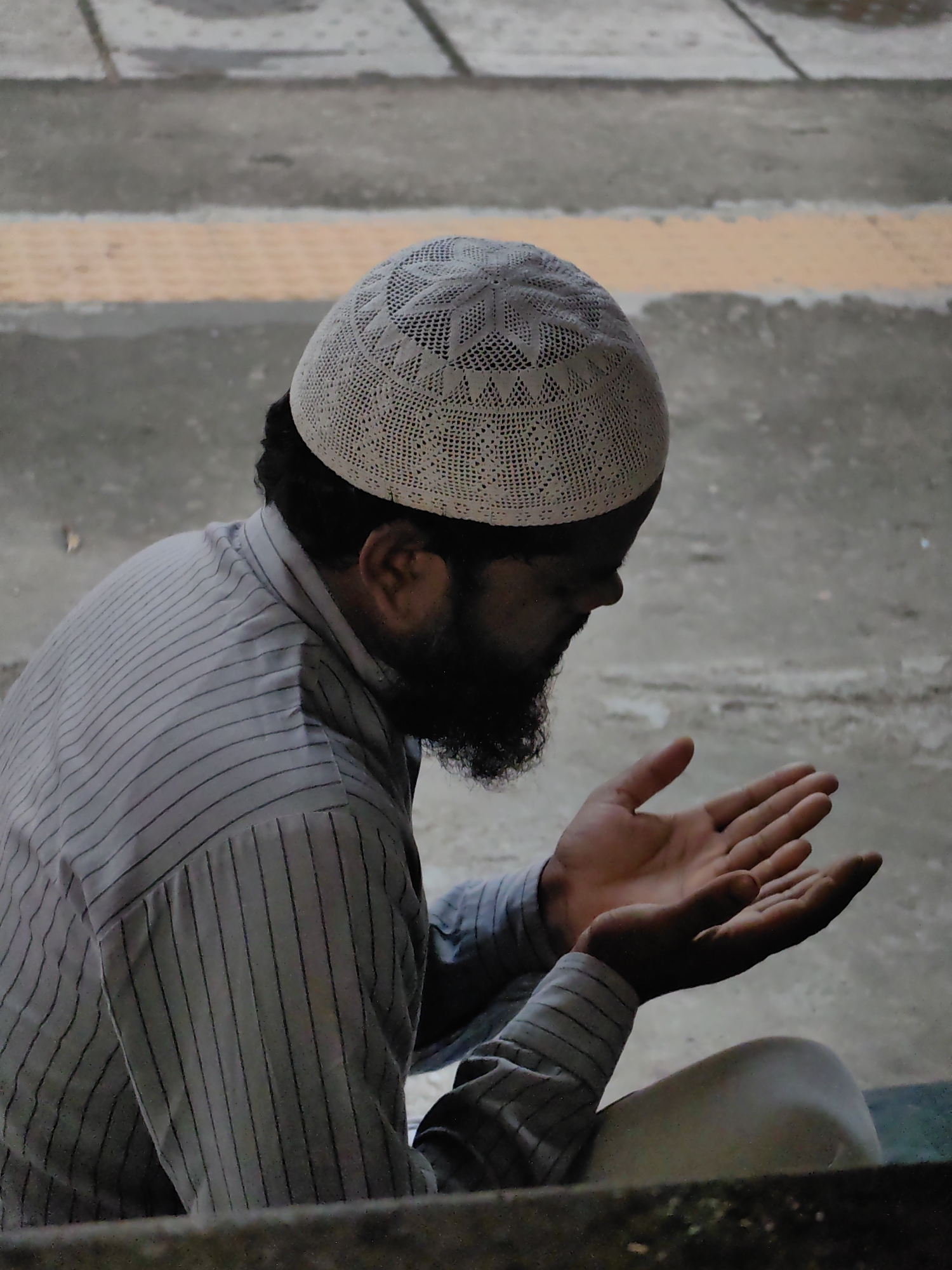A person is shown in profile, wearing a white kufi and a striped shirt, with their hands held open in prayer. They have a beard and are looking downwards. The background is a textured concrete surface with a yellow stripe. The overall scene appears to be outdoors, possibly in a public space within the city of Varanasi, India, given the context provided.
