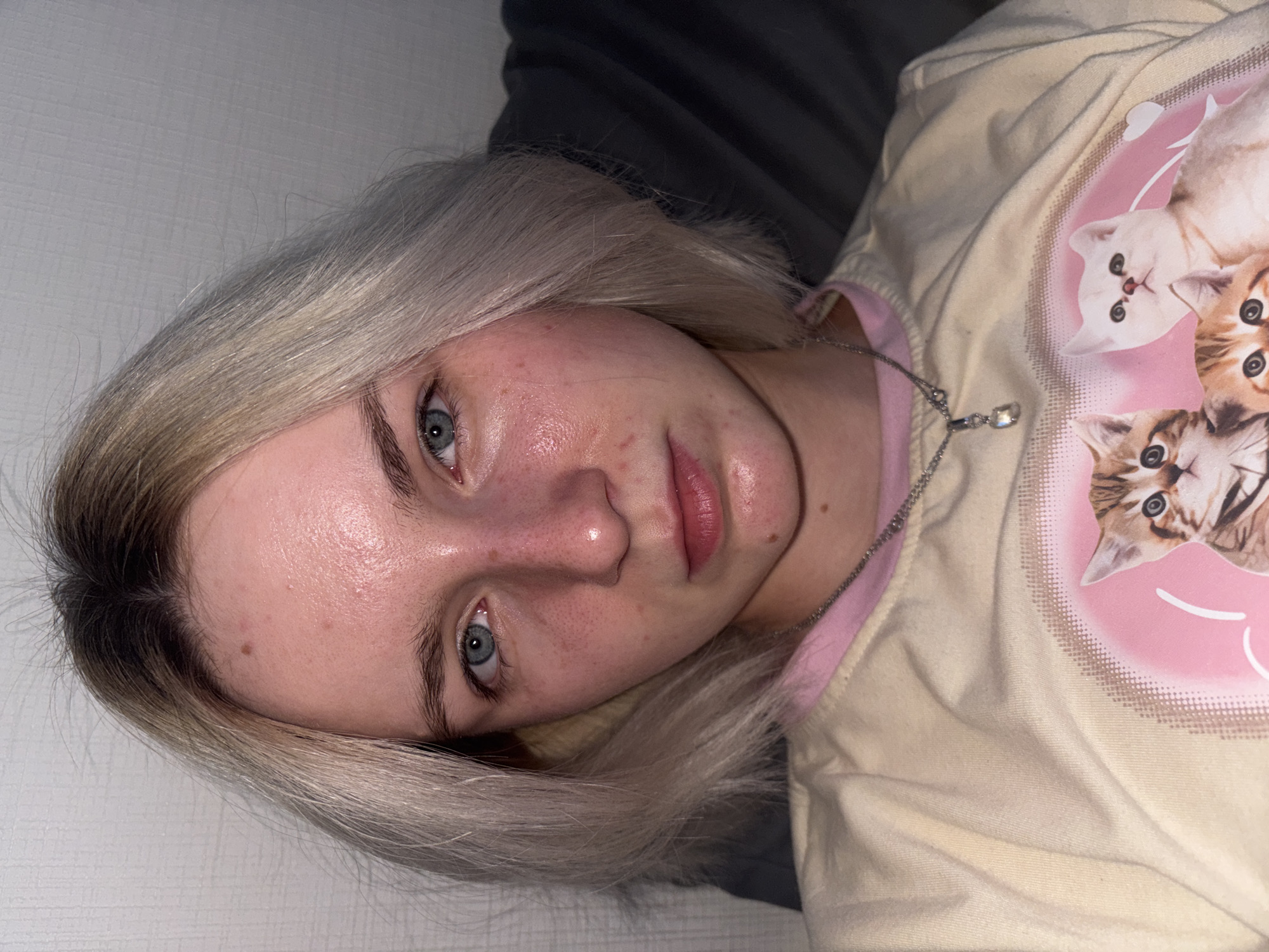 A close-up shot shows a woman lying down. She has fair skin, blue eyes, and blonde hair with dark roots. She is wearing a light-colored T-shirt with a pink-and-white graphic featuring kittens. A silver necklace is visible around her neck. The background consists of a gray surface and a white surface. No information in the image indicates the city of Vsevolozhsk, Russia.