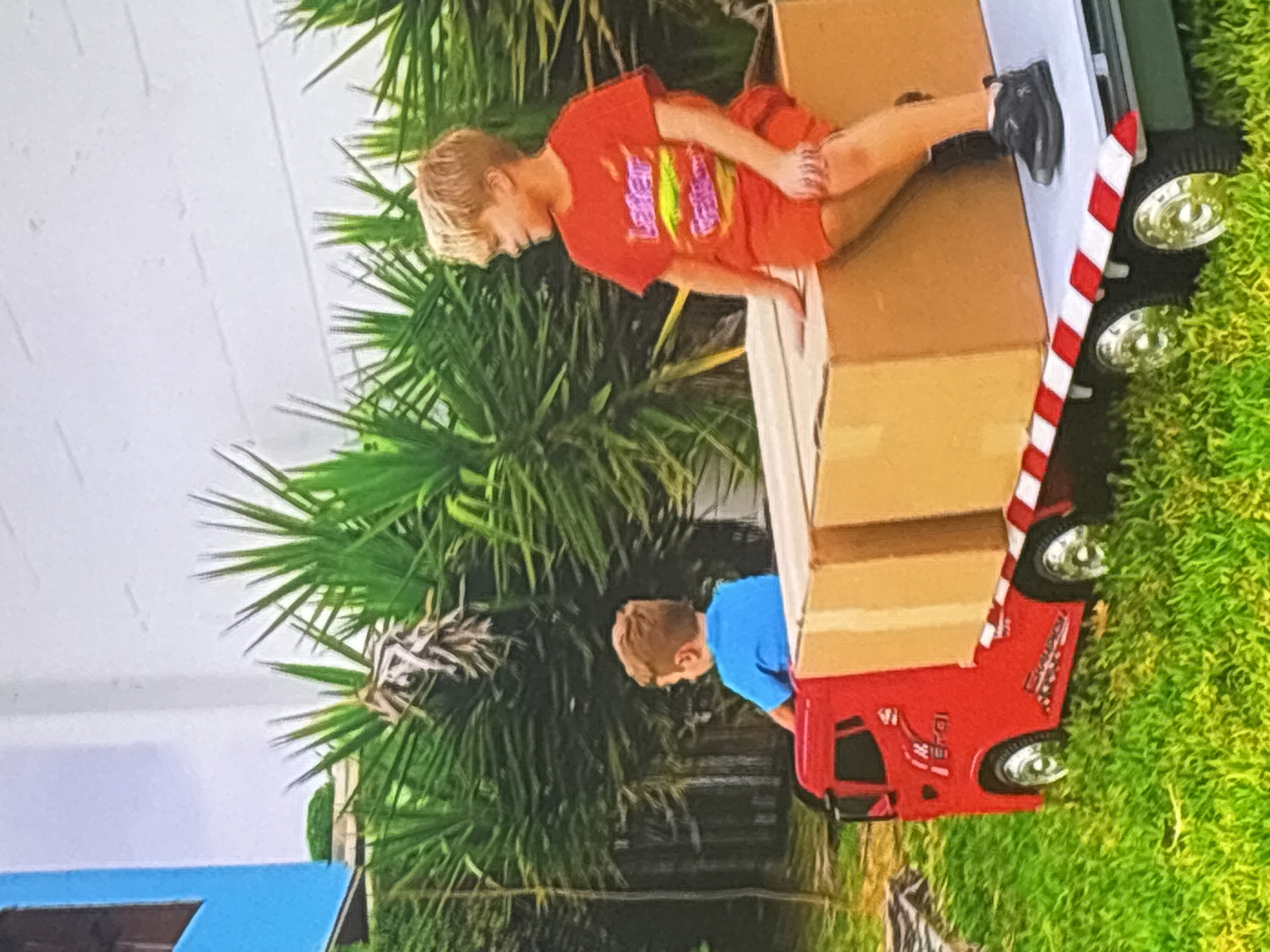 Two young boys are playing with a large toy truck in a grassy area. The truck is red and white, with a flatbed loaded with two cardboard boxes. One boy, wearing a blue t-shirt, sits in the driver's seat of the truck. The other boy, wearing an orange t-shirt and shorts, sits on top of one of the boxes. In the background, there are palm trees and a white building. The scene appears to be a sunny day.