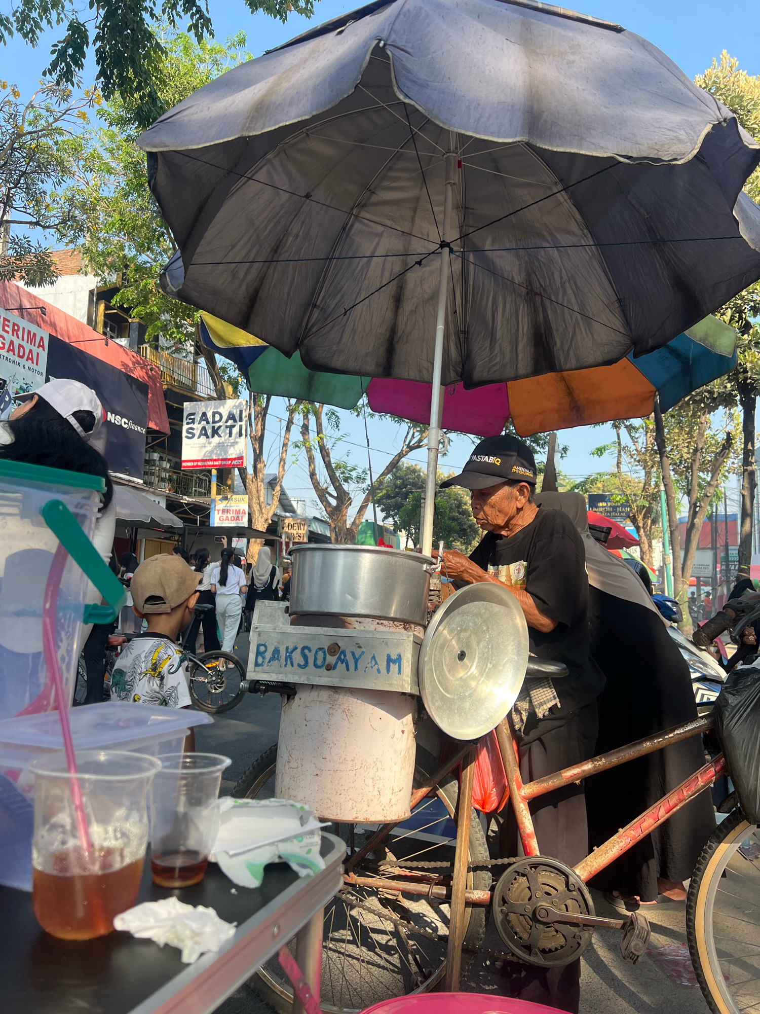 The image depicts a street scene in Kecamatan Pati, Indonesia, likely a market or roadside vendor area. The focal point is an elderly man, wearing a black t-shirt and a black baseball cap with the word "FASTABIQ" and a logo, operating a food cart that appears to be attached to a bicycle. He is preparing food, possibly bakso (Indonesian meatball soup), as indicated by the text "BAKSO AYAM" on the cart. A large, multi-colored umbrella, predominantly gray and with some yellow and blue sections, shades the vendor and his cart from the sun.
In the foreground, a young boy wearing a patterned t-shirt and a light-colored baseball cap is seated, facing away from the camera, seemingly observing the vendor. In front of him, a table holds two plastic cups, one with a pink straw and a brown liquid, and another partially filled with a similar liquid. There are also some crumpled white napkins on the table.
Behind the vendor, a woman wearing a hijab and a black outfit is partially visible. Other people are seen in the background, suggesting a bustling environment. Signs for businesses like "ERIMA GADAI" and "GADAI SAKTI" are visible on buildings in the background, indicating a commercial area. The presence of trees and a clear blue sky suggests it is daytime. The overall scene captures a slice of everyday life at a street food stall in Indonesia.