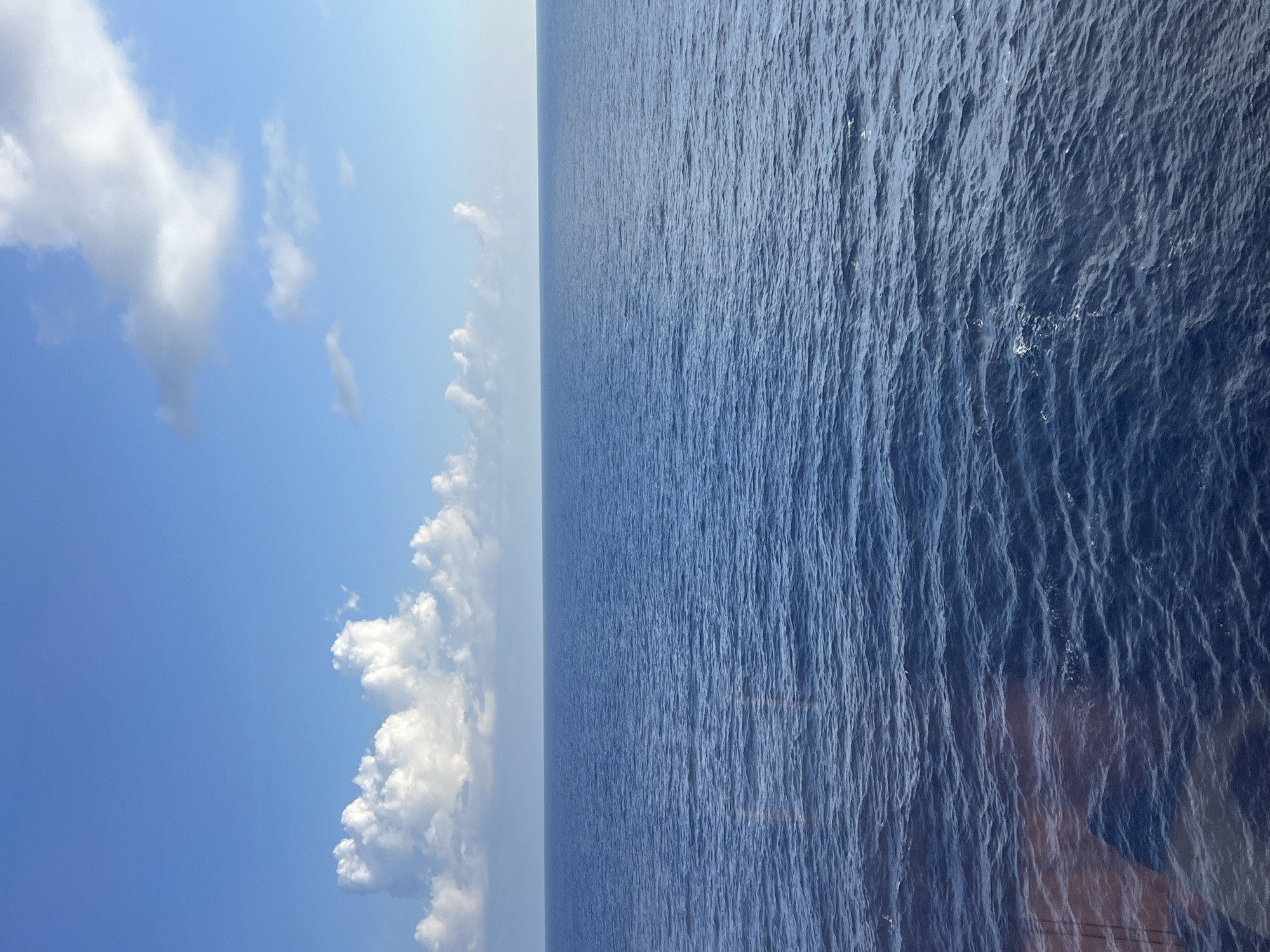 The image depicts a vast expanse of deep blue ocean meeting a clear sky with scattered white clouds. The horizon line is sharp, dividing the ocean and sky. Gentle waves ripple across the surface of the water, reflecting the sunlight. In the lower left corner, a subtle reflection or glare from a windowpane is visible, hinting at the viewpoint from an enclosed space, possibly a ship or building. The scene is tranquil and vast, with no immediate signs of human activity or specific landmarks.