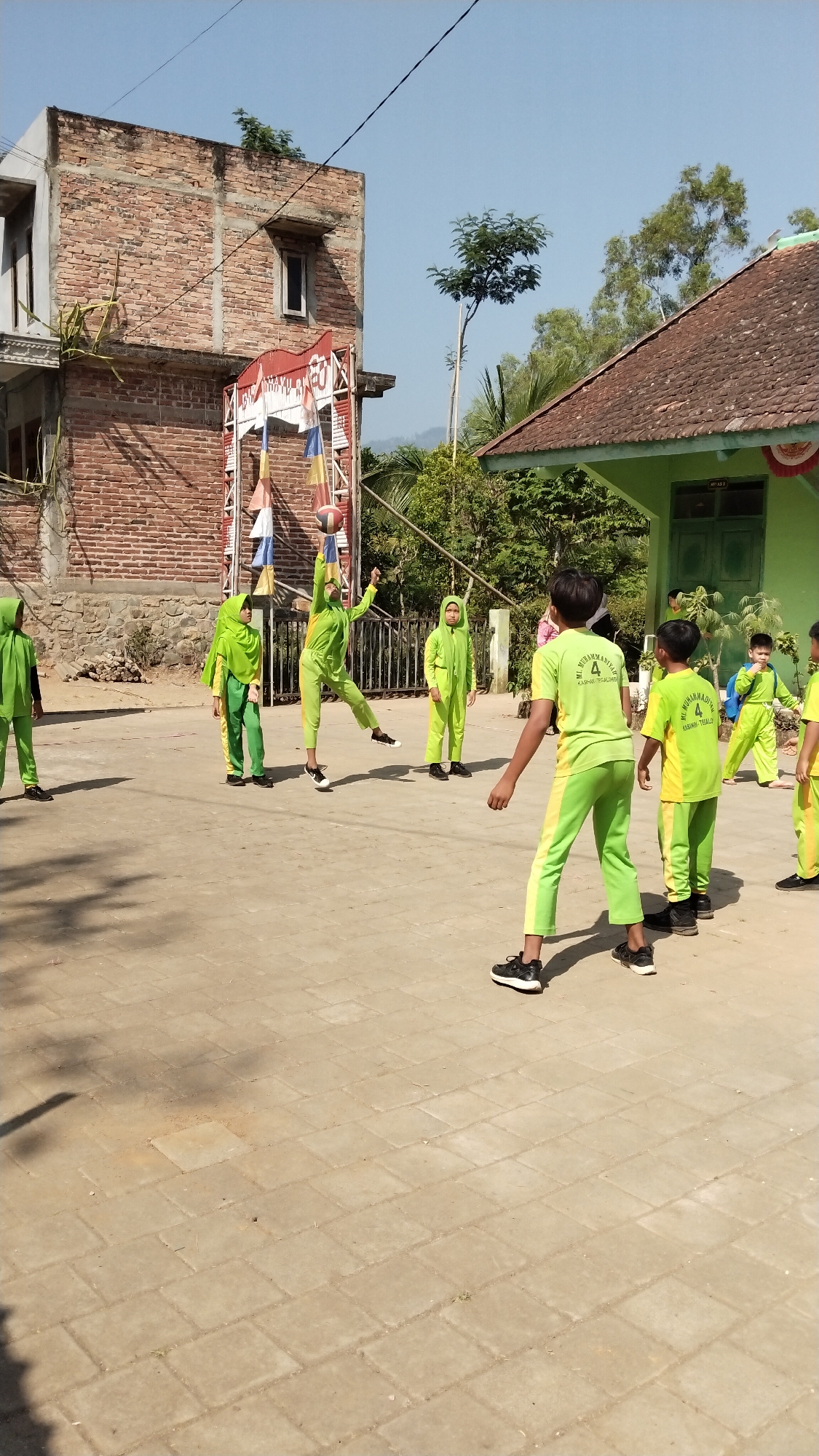 A group of children in bright green uniforms are playing with a basketball outside a building in Tegalombo, Indonesia. The scene is set on a sunny day, with a clear blue sky overhead. The children appear to be engaged in a game of volleyball or a similar sport, with one child in mid-air jumping to hit the ball. The background shows a brick building, a green-roofed structure, and lush green trees, suggesting a rural or semi-rural setting. The ground is paved with light-colored rectangular tiles.