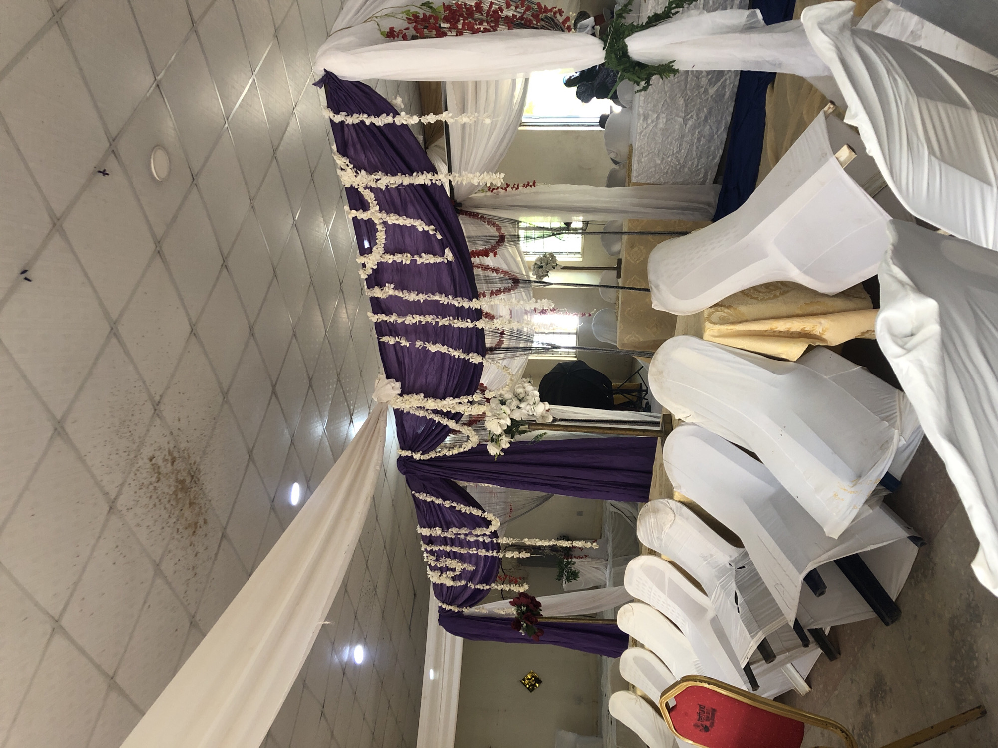 The image depicts an interior space, likely a venue in Akwanga, Nigeria, prepared for an event. The scene focuses on decorations, primarily white and purple fabric drapes adorned with white floral garlands, creating a ceremonial archway. Rows of chairs covered with white fabric are arranged in the foreground. The ceiling appears to be tiled, with some stains or imperfections visible. One chair has a red back, providing a splash of color among the predominantly white chair coverings. There is no specific human activity visible. The room seems to be in a state of preparation, not yet occupied by guests.