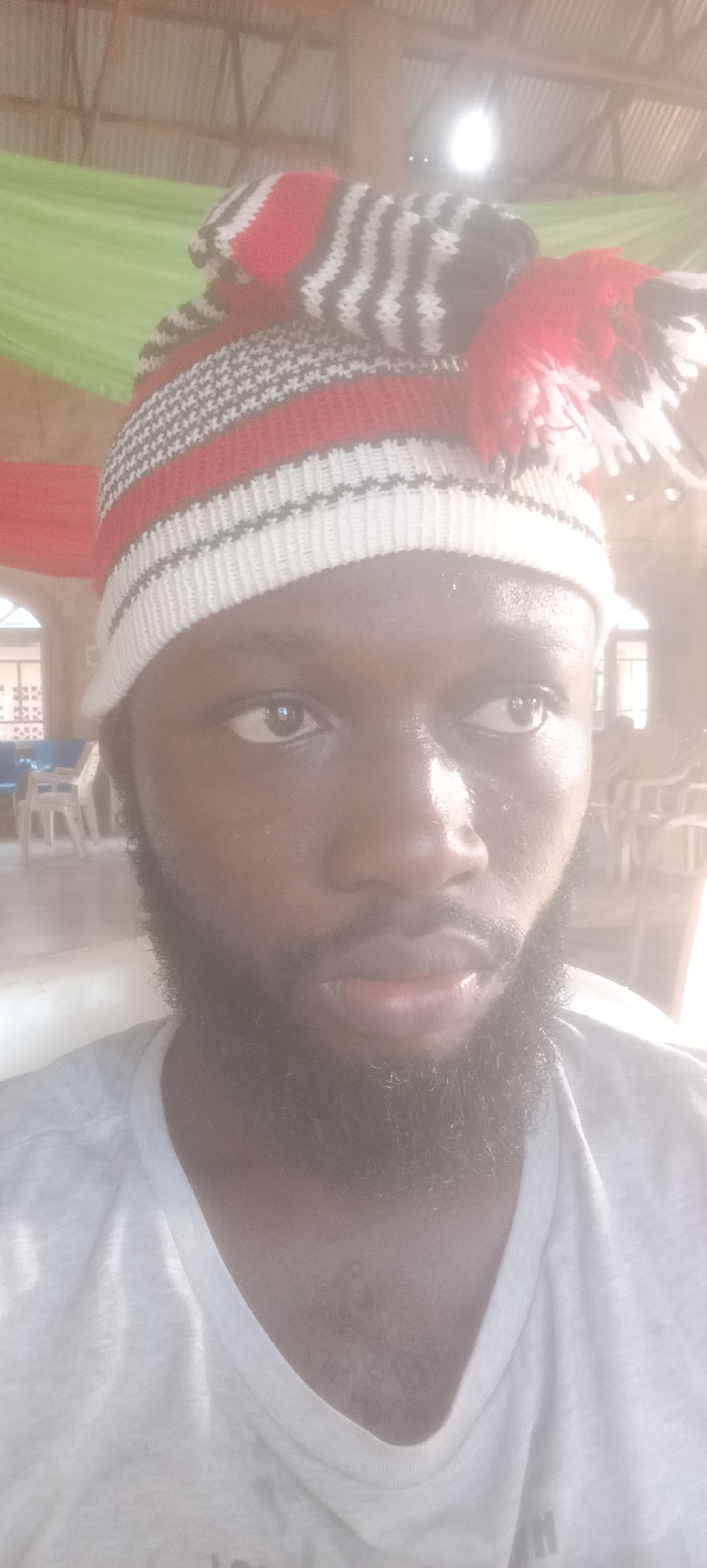 A man with dark skin and a full beard wears a white, red, and black striped knit cap with a pom-pom. He is looking off to the side with a serious expression. He is wearing a light grey t-shirt. The background appears to be an indoor space with tables and chairs. A green and red fabric drape is visible in the upper left corner. The location is likely in Bende, Nigeria.