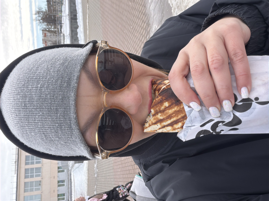 An adult individual is depicted in a close-up, chest-up outdoor shot within an urban pedestrian area of Chaikovsky, Russia. The individual wears a black hooded jacket, a grey knit beanie, and amber-framed sunglasses with dark lenses. The subject is actively biting into a grilled wrap, possibly a shawarma, held in both hands. The wrap is partially covered in white paper with black stylized designs, and a portion of its filling is visible where it has been bitten. The individual's visible left hand shows short, square-shaped fingernails painted with white iridescent polish. In the background, a light-colored, multi-story building with a flat roof and multiple rectangular windows is situated to the left. Further right in the background are bare trees, distant urban structures, and scattered patches of snow. The ground is paved with reddish-brown interlocking bricks, partially covered by melting snow and slush. A blurred partial figure of another person, carrying an object with colorful embellishments, is visible in the left midground. The sky is overcast.