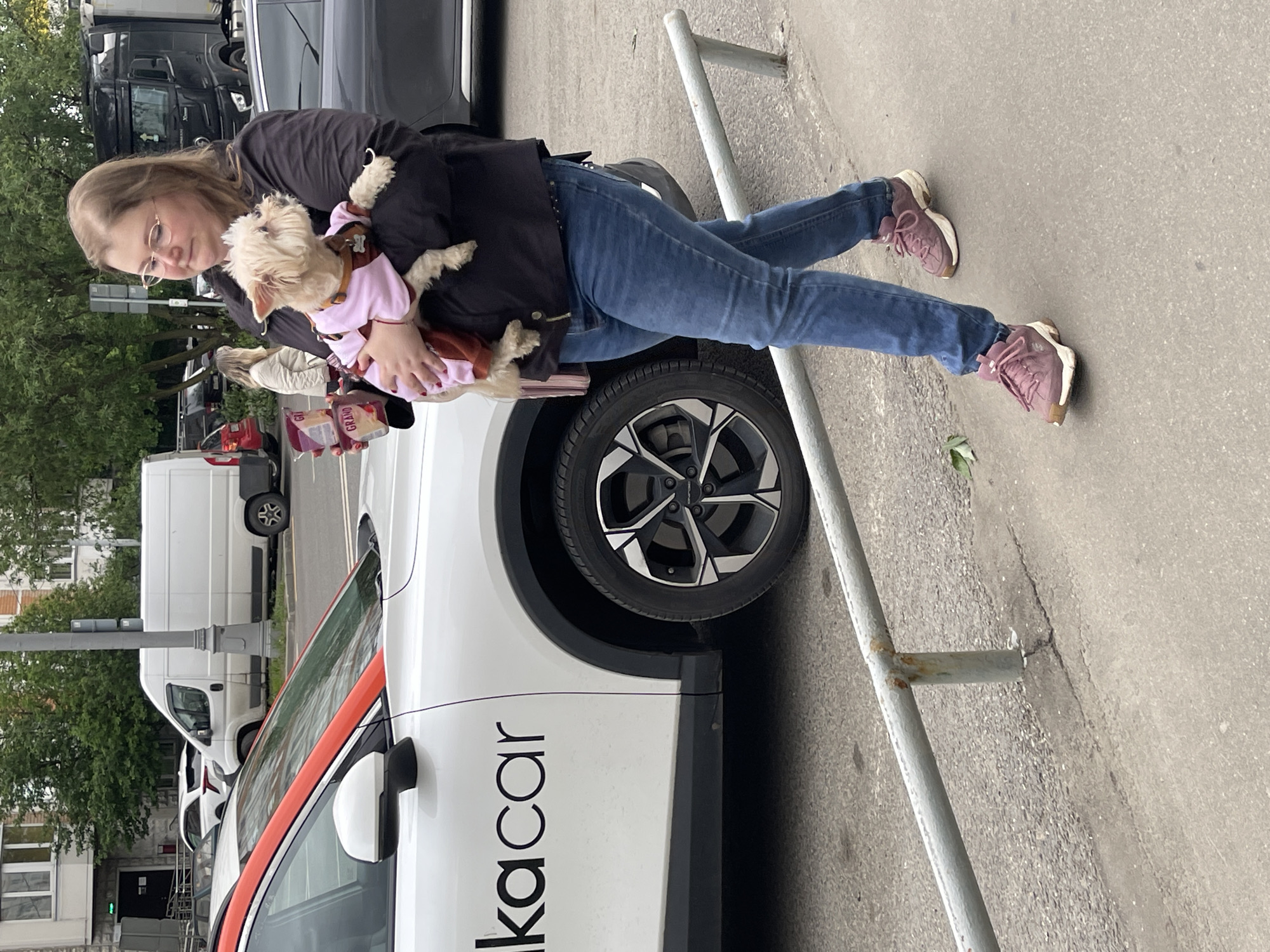 A woman is holding a small dog in a pink outfit in her arms. She is wearing glasses, a black jacket, and blue jeans with pink sneakers. She is standing next to a white car with the word "лкаcar" written in black letters on the side. The car has an orange trim around the side mirror. A metal parking barrier is next to the car. In the background, there are other cars and buildings, suggesting a city street. The location is most likely Moscow, Russia, due to the "лкаcar" branding, which is a car-sharing service in Russia.