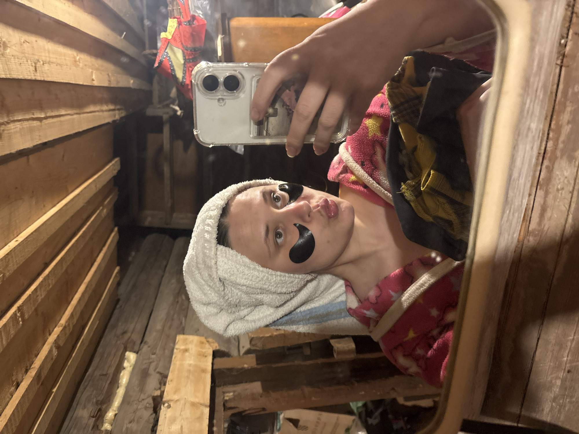 A woman takes a selfie in a mirror. She wears a towel on her head and eye masks under her eyes, and a pink robe with white stars. She holds a phone with a clear case. The reflection reveals the setting is a rustic, unfinished room or storage area with exposed wooden beams and planks along the walls. The location of Nizhnyaya Chermoda, Russia is not explicitly visible in the image, therefore I am unable to verify.