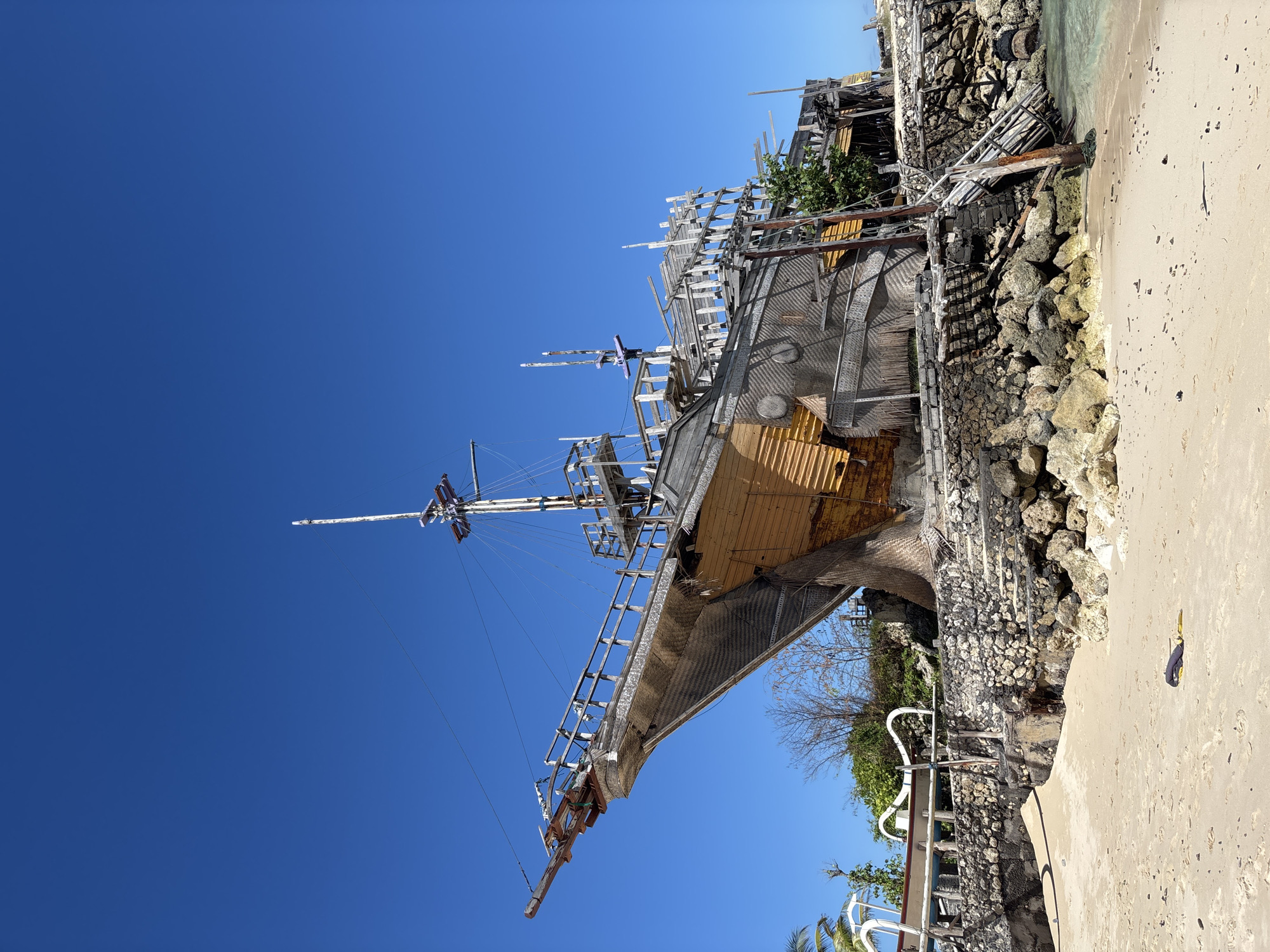 The image captures a partly decayed, wooden pirate ship replica, situated on a sandy beach adjacent to a rocky outcrop. The ship's hull is a mix of grey and yellow hues, with incomplete wooden framework suggesting disrepair or ongoing construction. The masts are present but lack sails. The beach is light-colored, and the sky is a clear, vibrant blue. The location is inferred to be in Kuta Selatan, Indonesia. The photo is taken from a low angle, looking upwards towards the ship and the sky. The image captures no individuals, but there are metal handrails on the rocks and other assorted debris surrounding the ship. The ocean water is present but only a small amount is visible.