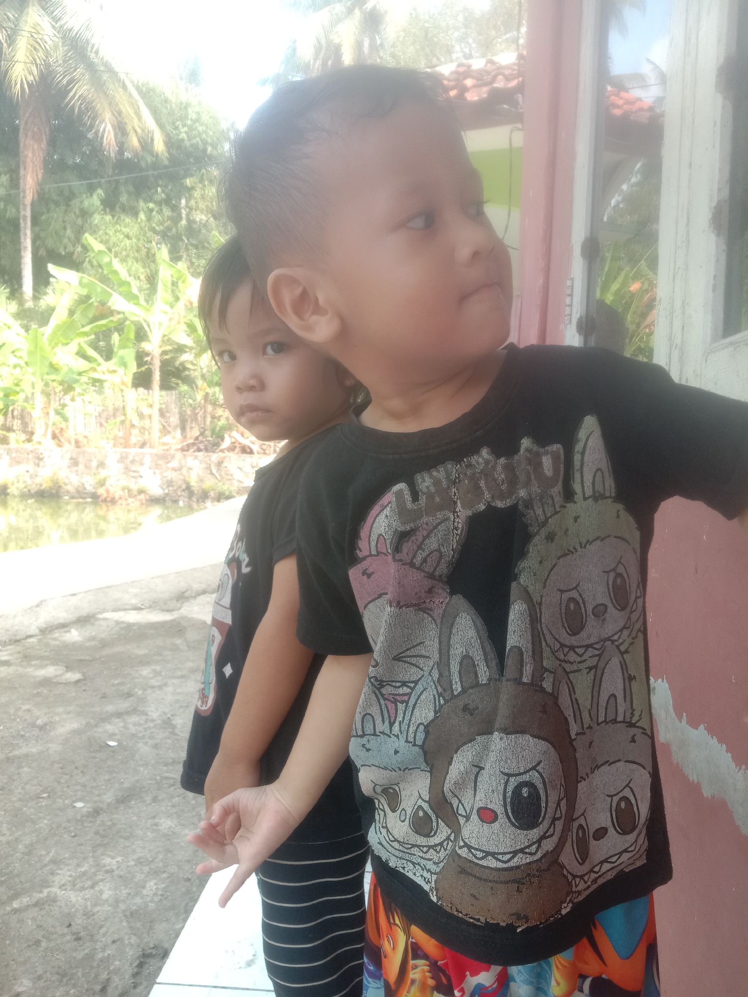 Two young children are posing for a photograph outdoors, likely in a residential area of Bojong, Indonesia. The child in the foreground, who appears to be older, is wearing a black t-shirt with a graphic print of cartoon characters, possibly bunnies. The child is looking to their right and slightly away from the camera. Behind them, a younger child is peeking out from behind their shoulder, also looking towards the camera with a curious expression. This child is wearing a black and white striped outfit. The background suggests a tropical environment with palm trees and lush greenery. The setting appears to be close to a body of water, possibly a canal or pond, and there is a building with a pink and white facade to the right of the children. The overall lighting suggests it is daytime, possibly late morning or early afternoon.