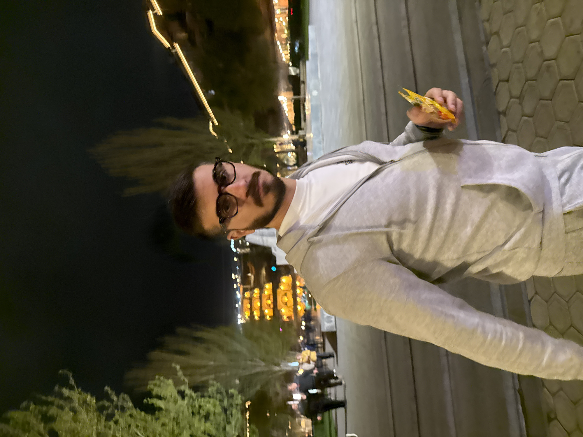 A man with dark hair and a beard, wearing glasses, a white T-shirt, and a gray hoodie, stands outdoors at night. He's holding a packet of chips in his right hand. The background reveals city lights, greenery, and what appears to be a waterfront or canal setting, suggesting the city of Yerevan, Armenia. The ground is paved with hexagonal tiles.