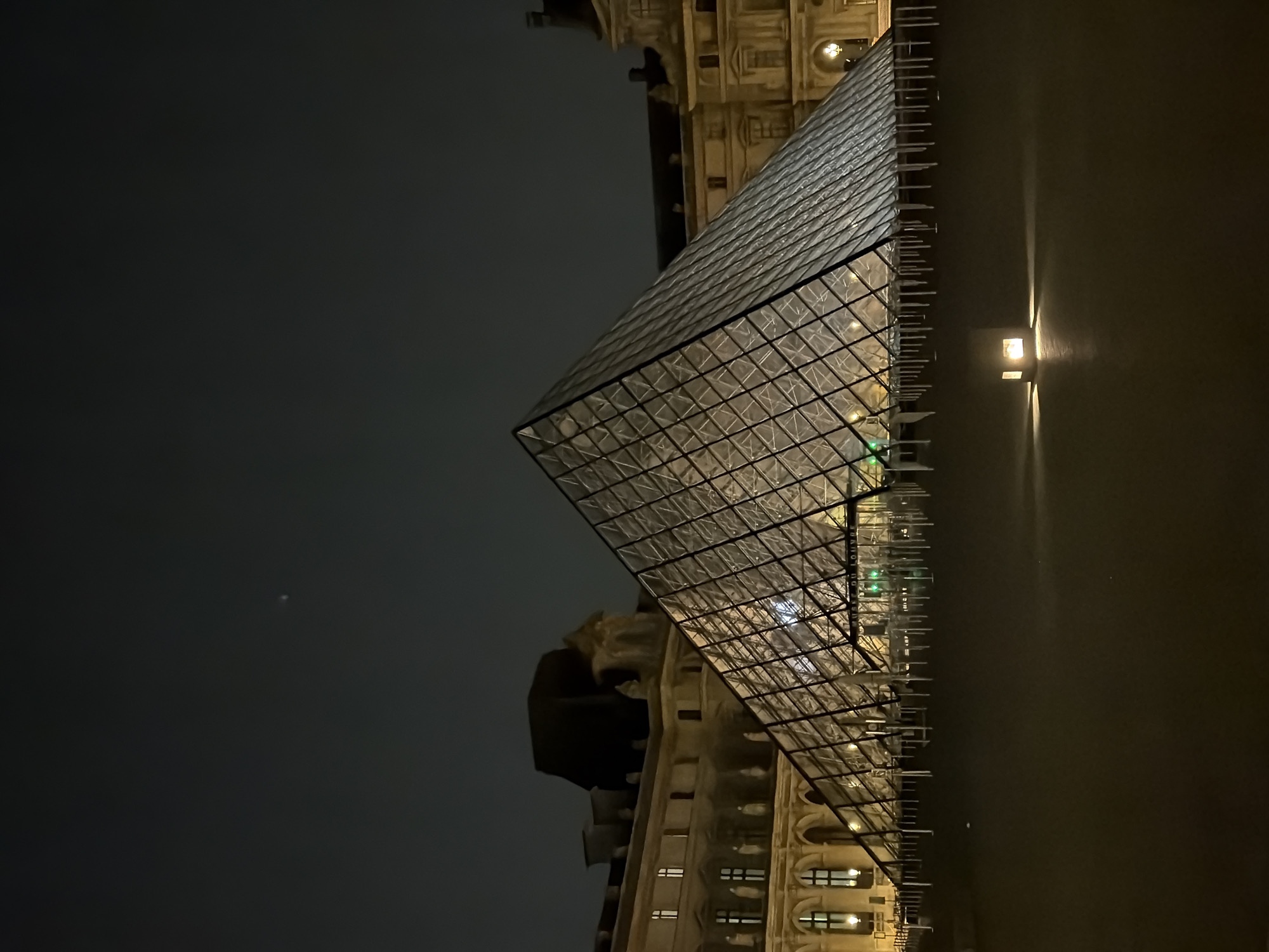 The image shows the Louvre Pyramid in Paris, France, at night. The pyramid's glass panels are illuminated, contrasting with the dark sky. Part of the Louvre Museum's architecture is visible behind the pyramid. A light source is visible in the background, reflecting on the ground. The moon is faintly visible in the upper left of the frame.