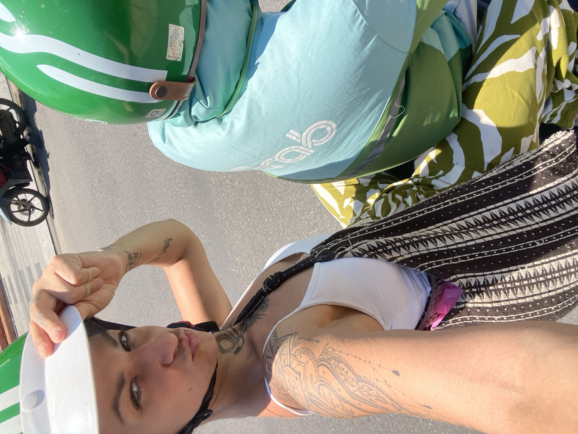 A woman, wearing a white helmet and a white tank top, is taking a selfie on a scooter. She has several tattoos on her arm and neck. She is wearing a patterned fabric sling bag over her shoulder. Behind her, another person wearing a green helmet and a light blue jacket with the Grab logo is sitting on the scooter. The scooter is on a paved road. In the background, a motorbike is visible on the road. The scene appears to be in Phu Thuy Ward, Vietnam.