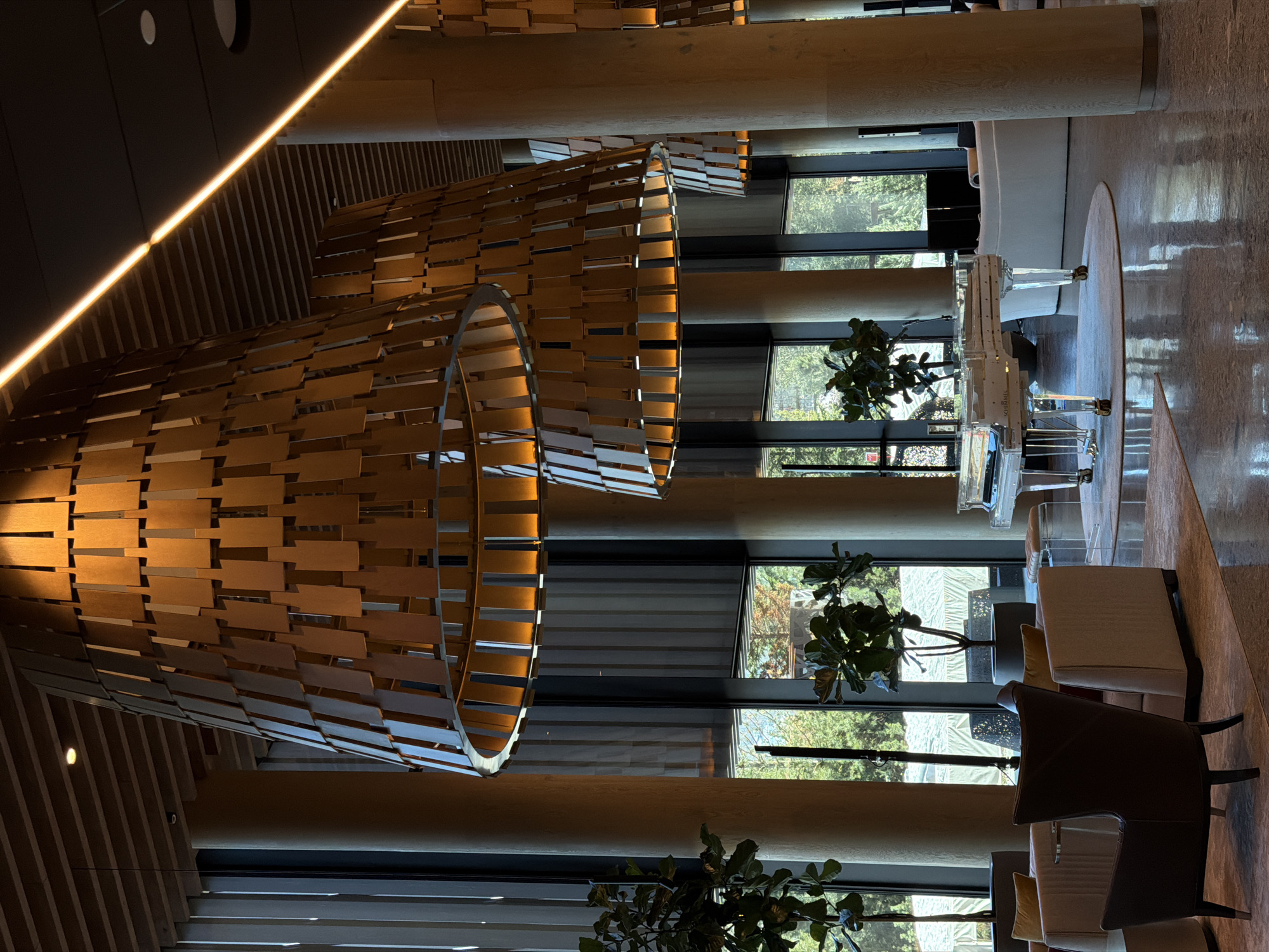 The image captures a bright and modern interior space, possibly a lobby or lounge. The most striking features are the large, cylindrical wooden art installations hanging from the ceiling, appearing as if constructed from numerous small, rectangular wooden tiles. A light fixture runs along the edge of the ceiling. Tall, round columns support the structure, and large windows reveal an exterior filled with greenery, suggesting a location with significant tree cover.
The furniture present includes chairs and sofas, some upholstered in light colors, positioned to create a seating area. A transparent piano, perhaps made of acrylic or glass, is centrally located. The floor is polished, reflecting light and contributing to the overall brightness. Potted plants add touches of nature to the interior. There are no people visible in the frame. The scene appears tranquil and upscale.
