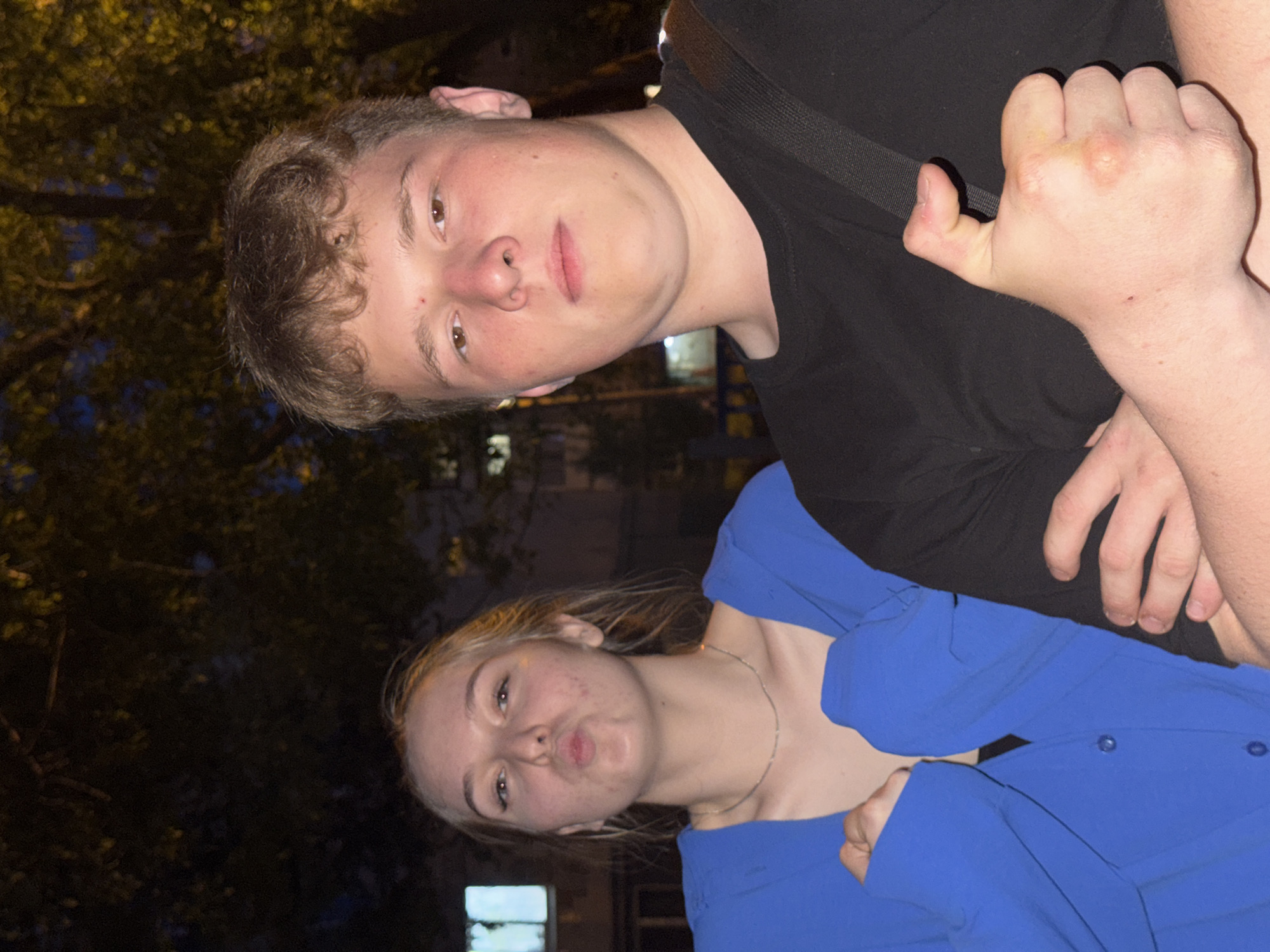 Two people are visible in the image. A young man with short brown hair is wearing a black t-shirt and a black strap. He has a fair complexion and is looking at the camera with a neutral expression. He is holding his hand in a fist. Beside him, a young woman with blond hair is wearing a blue shirt. She is making a kissing face. The background appears to be a dimly lit outdoor scene, with trees and a building with a window visible. It is reported the location is in the city of Perm, Russia.