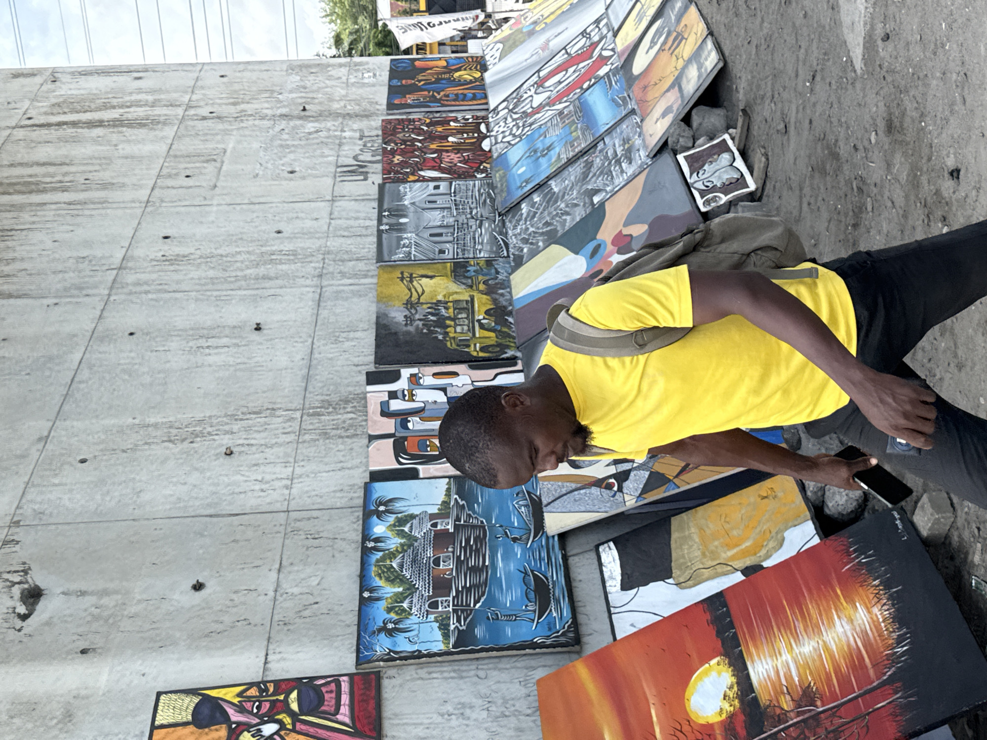 An outdoor scene in Lekki, Nigeria, captures a man displaying a vibrant collection of paintings. The image is taken from a slightly elevated perspective, looking down at the street-level art display.
The primary subject is a dark-skinned man, seen from the waist up, wearing a bright yellow short-sleeved t-shirt and dark trousers, with a beige backpack slung over his shoulder. He has short dark hair and a beard and is looking down, holding a black smartphone in his right hand.
He is surrounded by numerous paintings of various sizes and styles, laid out on the dusty, gravelly ground and leaning against a large, grey concrete wall. The artworks depict diverse subjects including abstract portraits, tropical landscapes with houses and boats, urban scenes with buildings and vehicles (some in black and white, others colorful), abstract patterns, and a vivid sunset over a dark landscape. One small painting on the ground shows a pair of shoes.
The setting appears to be an informal art market or a street vendor's stall, possibly under an overpass given the imposing concrete structure. The concrete wall features visible formwork lines, small holes, and some dirt smudges. Partially visible text on the wall appears to read "UNCREAT" or "UNCPERT". The lighting is bright, suggesting daytime, and the sky above the concrete structure shows some power lines or cables. The overall impression is one of a bustling, creative urban environment.
