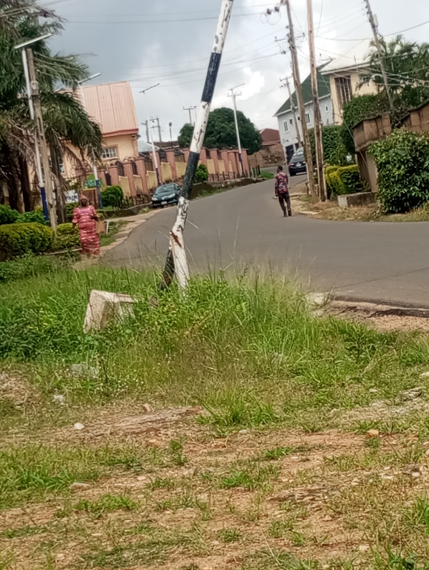 This image depicts a street scene in Jos, Nigeria. In the foreground, there is overgrown grass and patchy soil with a rough, beige concrete object, possibly a broken part of a structure or signage, lying on its side. A black and white striped pole, part of a barrier or gate, stands vertically on the right side of the image.
The middle ground shows a paved road curving to the left. On the left side of the road, a person is walking away from the camera, wearing a red and white patterned dress. Further down the road on the left, a dark-colored car is parked or driving. A cluster of buildings with reddish-pink and pale yellow facades, surrounded by decorative fences and manicured green bushes, lines the left side of the street. Palm trees and other foliage are visible, suggesting a tropical or subtropical climate.
On the right side of the road, more green bushes and a multi-story building with white walls and a green roof are visible. A second person, wearing a dark patterned shirt and trousers, is walking away from the camera on the right side of the road, appearing to carry a bag or backpack. Utility poles with wires are present along the street, indicating the presence of electrical infrastructure. The sky is overcast, suggesting a cloudy or possibly rainy day. The overall impression is a residential or semi-commercial street in a well-established neighborhood.