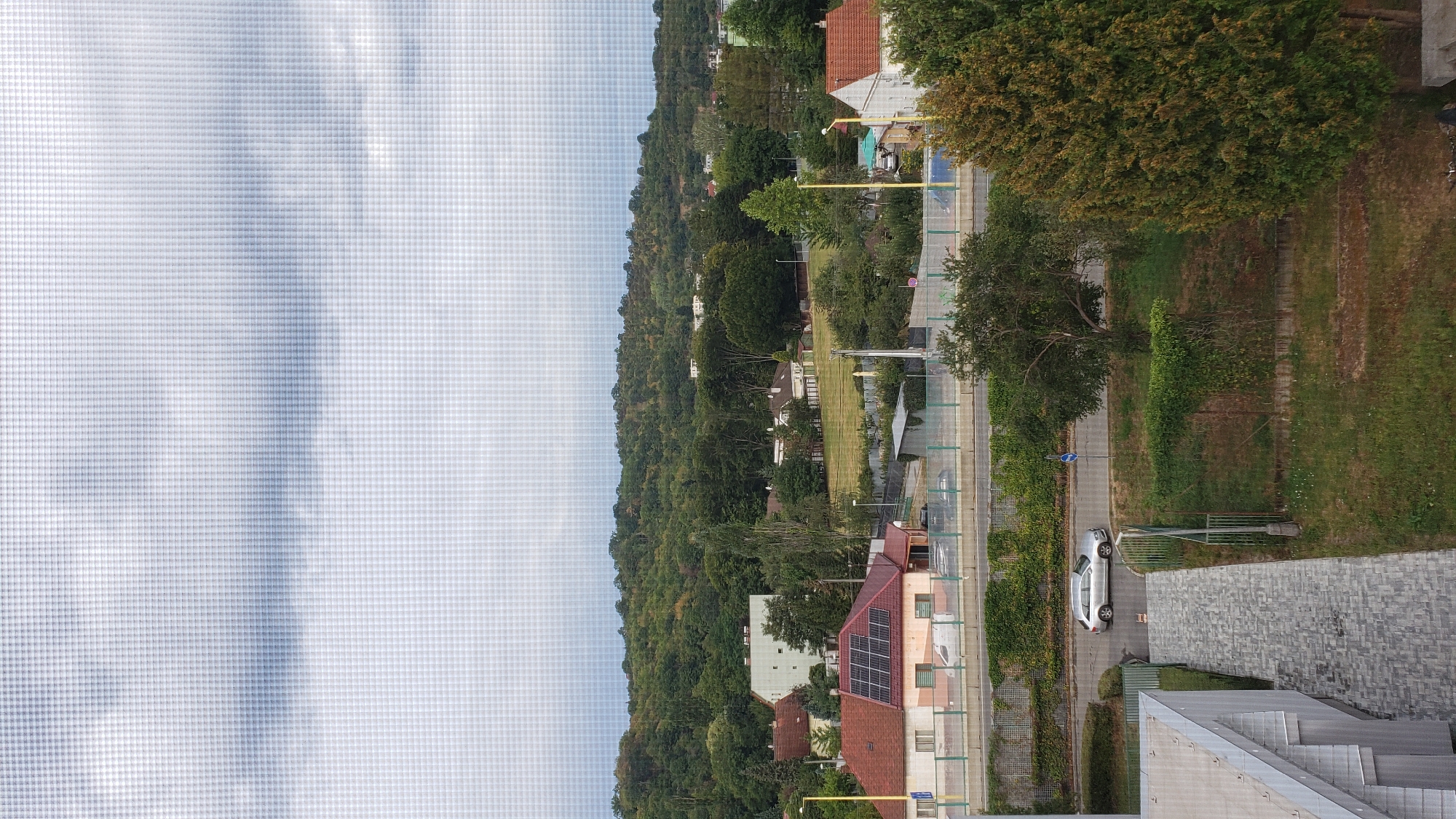 This is a medium-shot, outdoor, vertical, eye-level photograph taken through a textured window screen, showing a suburban landscape with a view of a tree-lined hill in the background. The sky is overcast and pale grey. In the mid-ground, a residential street runs horizontally across the frame, with a silver sedan parked on the right side of the road. Several houses with red tile roofs are visible, one with solar panels. A light-colored multi-story building is situated further back, behind the houses. The foreground features a portion of a modern building with a sloped roof and a grey stone-tiled walkway leading towards the street. The scene appears to be in a residential area, likely in Prešov, Slovakia, characterized by a mix of houses, greenery, and infrastructure.
