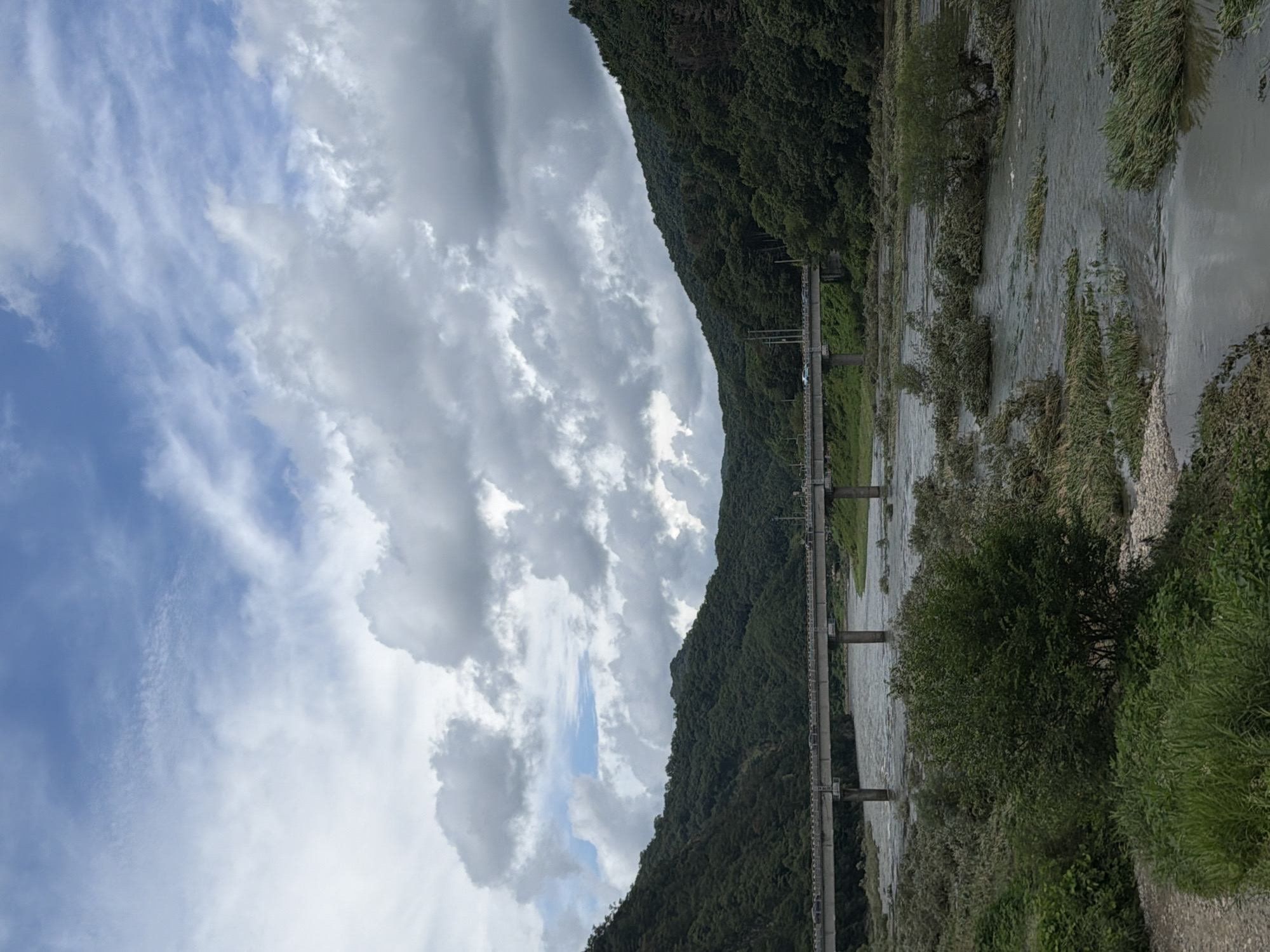 The image depicts a wide river flowing through a valley with lush green hills on either side. A long, concrete bridge spans the river, with several vehicles visible crossing it. The sky is overcast with dramatic, grey clouds interspersed with patches of blue. The river appears to be flowing with some force, carrying debris and submerged vegetation. In the foreground, the riverbank is covered in dense green foliage and smaller bushes. The overall scene suggests a natural landscape with human infrastructure integrated into it, likely in a rural or semi-rural area. The location is identified as Seo-myeon, South Korea.