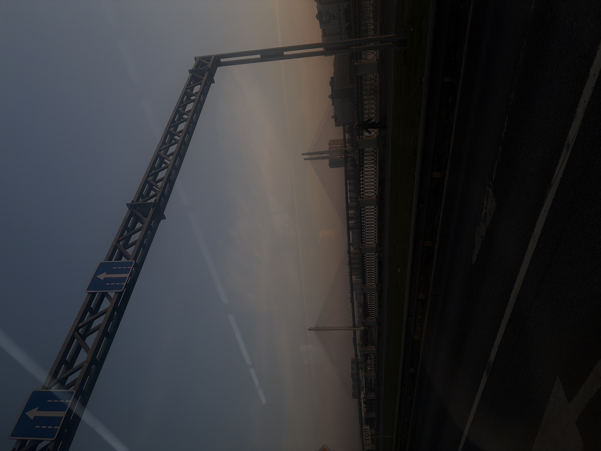 Here's a description of the image:
The photograph depicts the cityscape of Saint Petersburg, Russia, specifically featuring the Saint Petersburg bridge and a roadside traffic sign. The shot is taken from a low angle, likely from a vehicle.
In the foreground, there is a metal structure holding up traffic signs with blue arrows pointing left. The background shows a view of the city with a bridge and buildings. The sky is overcast, and the overall lighting is dim. There is also a person standing on the pedestrian walkway, near the ornate railing. To the right is the road with a white line in the center.