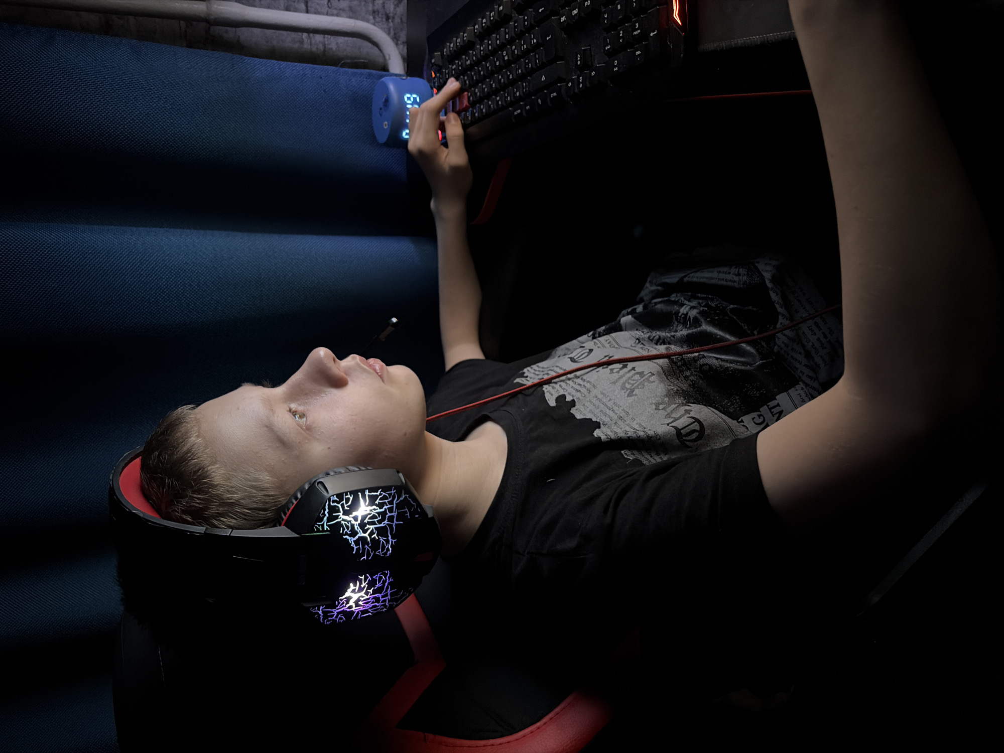 A young male individual is depicted seated indoors in the city of Chaikovsky, Russia, in a dimly lit environment, engaged with a computer setup. The subject, with short, light-colored hair, is shown in profile facing right, wearing a black short-sleeved t-shirt with a subtle graphic pattern. He wears over-ear gaming headphones that feature multi-colored, illuminated lightning-bolt designs on both earcups and a small microphone extending towards his mouth. His right arm is positioned over a dark surface, likely a desk, with his right hand resting on a black gaming keyboard which exhibits red backlighting on specific keys. A red key is depressed by his right index finger. Adjacent to the keyboard on the right, a small, blue cylindrical digital device displays "0:49." The subject is seated in a gaming chair with a visible red-accented backrest. The background consists of dark blue, textured curtains on the left and center, partially revealing a light-colored, possibly brick or concrete, wall with a vertical white pipe on the far right. The illumination primarily originates from the electronic devices and an unseen source to the left of the subject.