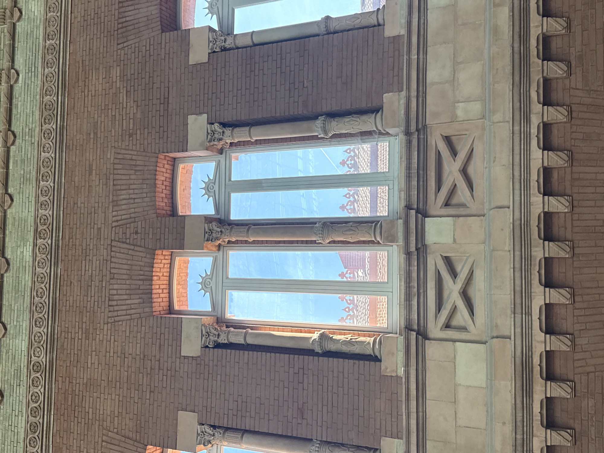 This media file presents a detailed, upward-looking view of an ornate building facade in Madrid, Spain. The structure is characterized by a rich interplay of reddish-brown brickwork and lighter-colored stone and decorative elements.
The central focus includes several tall, rectangular windows with white frames. Each window features a distinctive sunburst or starburst motif integrated into its upper glass pane. Through these windows, a bright, clear blue sky is visible, indicating sunny weather during the day, likely mid-morning or early afternoon.
Between the windows are decorative columns, crafted from a lighter material like stone or concrete, adorned with intricately carved capitals and bases. The surrounding brickwork exhibits varied patterns, including sections arranged in fan-like or radiating designs around the windows, adding to the architectural complexity.
To the right of the windows, a broader vertical section of the facade is constructed from lighter stone blocks, featuring a prominent rectangular panel with a bold 'X' shaped relief carving. Further right, a darker brick section displays a series of projecting architectural elements, possibly corbels or brackets, each with distinct geometric patterns. Along the top left of the image, a horizontal decorative band or frieze runs across the facade, showcasing repeating circular or floral medallion motifs.
The image is devoid of people or discernible activities, focusing solely on the intricate and static architectural details. No visible text is present within the captured scene.