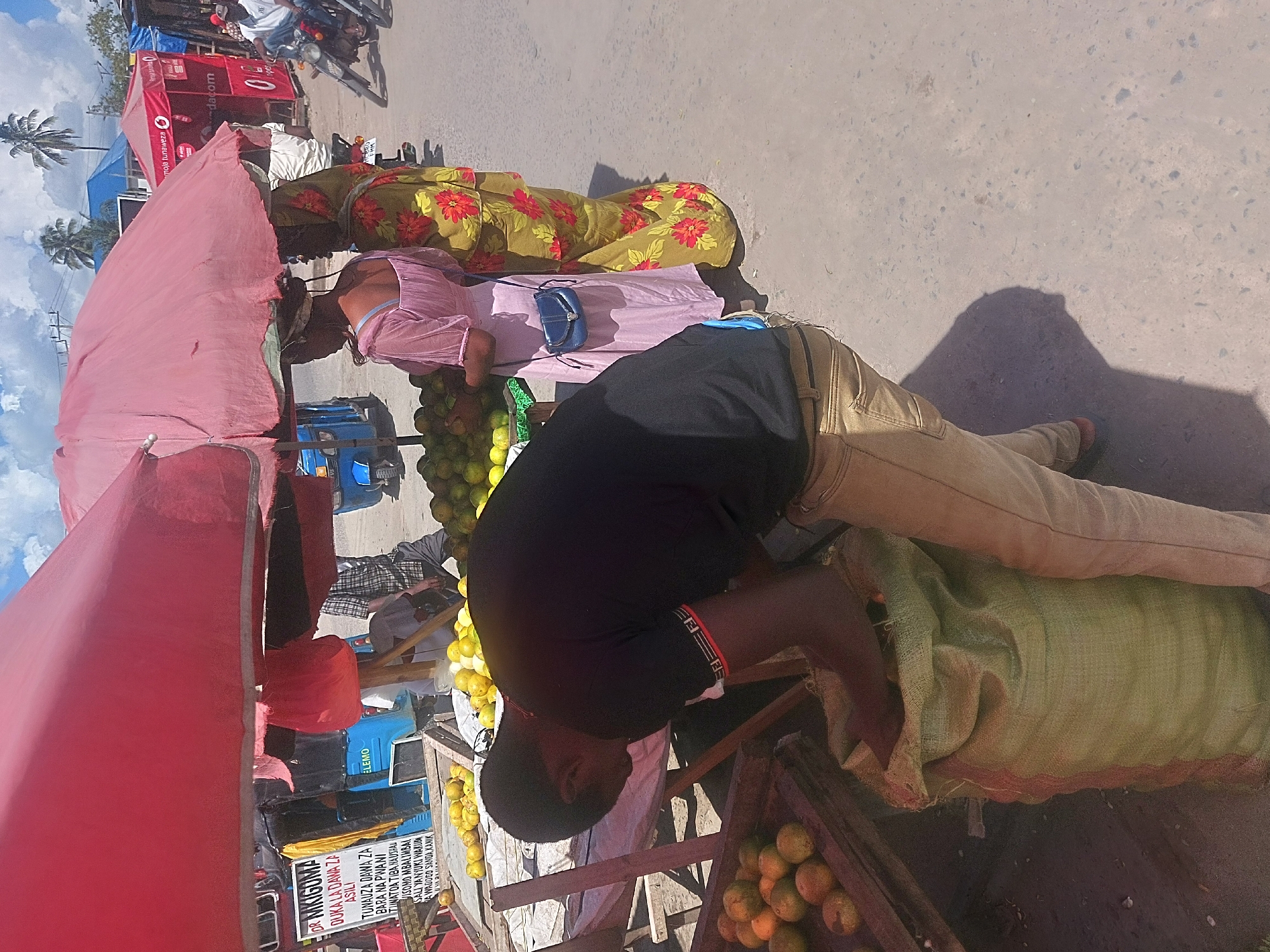 This image shows a street scene in Dar es Salaam, Tanzania, where vendors are selling produce. A man wearing a black shirt and tan pants leans over a large sack, possibly filled with fruits or vegetables. Next to him, a wooden crate contains oranges. Two women are also present, one wearing a pink dress with a small blue purse, and the other in a green and yellow floral dress. They appear to be working at the stall as well. The produce stand is set up under a pink parasol. In the background, there's a red Vodafone-branded stand, a blue truck, motorcycles, and a sign advertising "Dr. Mkigoma" as a seller of traditional medicine. The overall setting appears to be a bustling roadside market.