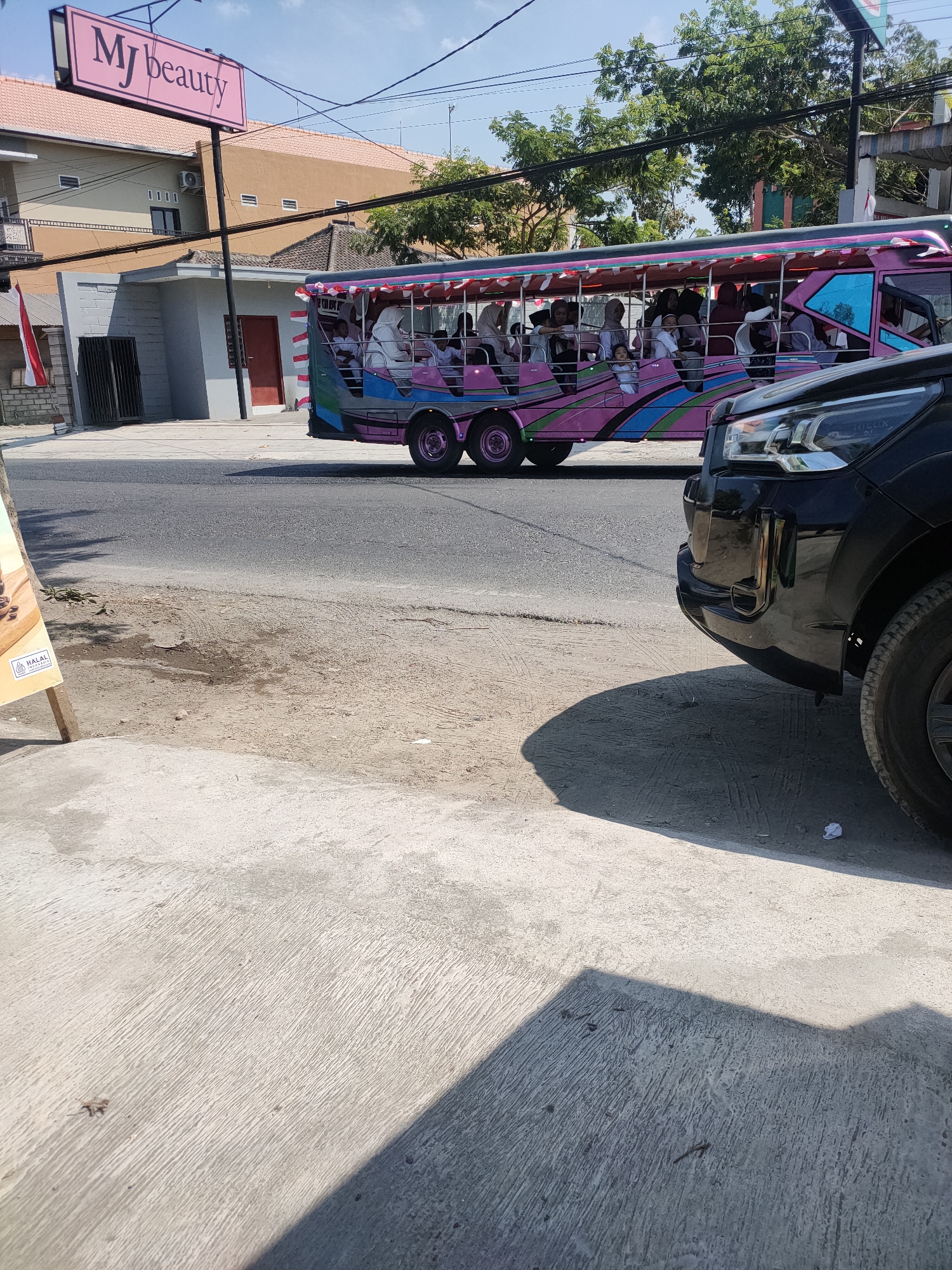 In Kanigoro, Indonesia, a vibrant, open-air bus, adorned with colorful decorations and a pink frame, transports a group of passengers. The bus, likely a form of local transport or tourist attraction, is seen on a paved road. In the foreground, a black car is parked, its front bumper and wheel visible. To the left of the image, a sign with the text "MJ beauty" in pink letters is prominently displayed, suggesting a commercial establishment nearby. The background consists of buildings and trees, under a clear blue sky. The scene depicts a typical day in this Indonesian city, with elements of daily life and local culture.