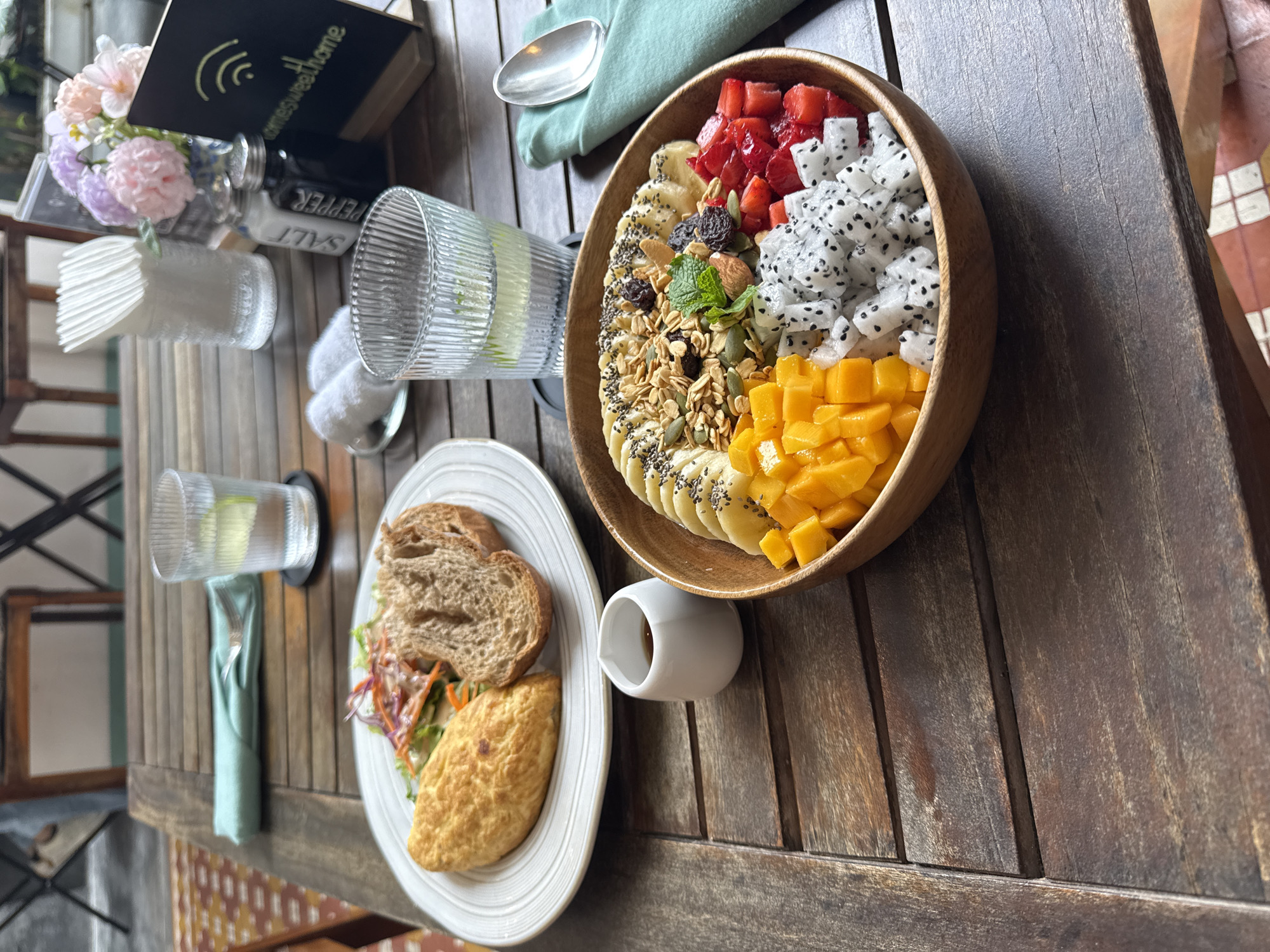 The image shows a table setting, presumably in a cafe, with a wooden table surface. There is a wooden bowl filled with fruit, granola, and seeds. The fruit selection includes strawberries, dragon fruit, mango, and banana. There is also a plate of food with sliced bread, an omelet, and some salad. Other objects on the table include a salt and pepper set, a vase of flowers, drinking glasses, a water pitcher, a napkin, a spoon, and a small white container. The location is likely Đà Nẵng, Vietnam, based on the overall aesthetic and assumed culinary style. There are no people visible in the image.
