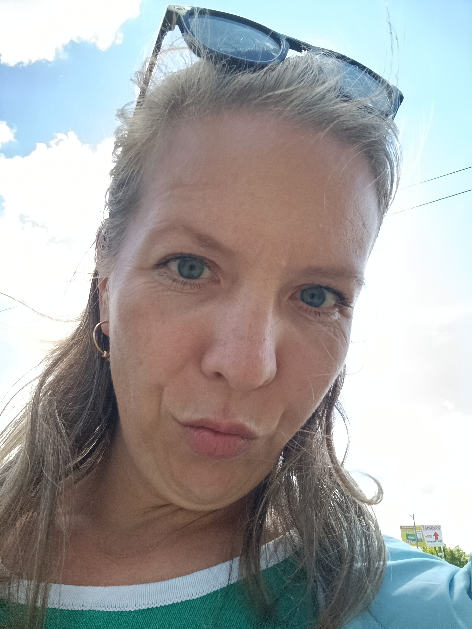 A selfie of a middle-aged woman with fair skin, blue eyes, and blonde hair is captured from a low angle against a bright, partly cloudy sky. She is wearing sunglasses pushed up on her head and gold hoop earrings. Her expression appears to be a pout. The woman is wearing a green and white shirt, with a piece of light blue fabric visible as well. In the background, partially obscured by greenery, is a sign in Russian located in the city of Chaikovsky, Russia. Power lines are visible near the top right.
