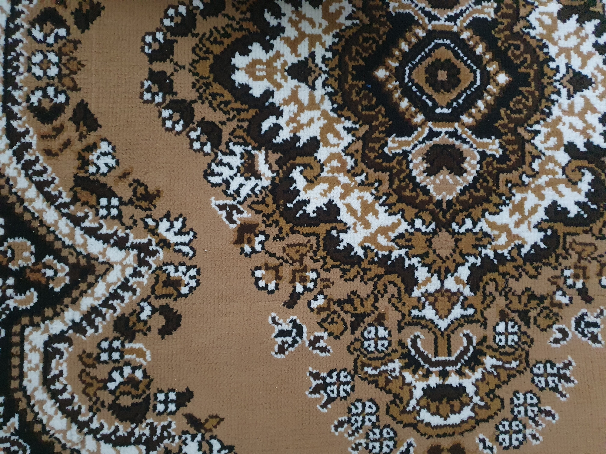 The image is a close-up, overhead view of a brown rug with a complex, repeating floral and geometric pattern. The pattern features intricate motifs in shades of brown, white, and black. The rug appears to be made of synthetic fibers, as suggested by the texture visible in the close-up crops. There are no people, animals, or other objects clearly depicted in the image, nor any specific environmental context beyond the rug itself. The location provided, Zubrești, Moldova, is not discernible from the visual information in the image.