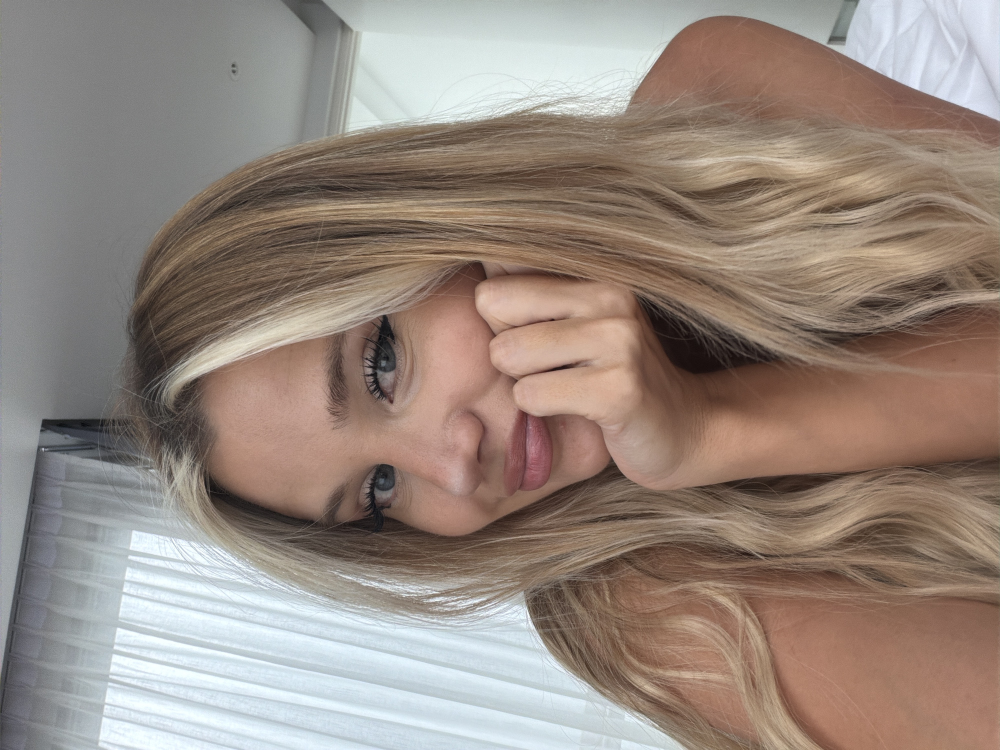 A medium shot photo captures a young woman with blonde hair, fair skin, and blue eyes. She is positioned in the center of the frame, resting her chin on her right hand. Her lips are slightly parted, and she is looking directly at the camera with a soft smile. She is wearing makeup, including mascara and eyeliner, and her hair is partially covering her left eye. The background consists of white curtains on the left and a lightly colored wall on the right, suggesting an indoor setting, potentially a bedroom, in Thep Krasatti, Thailand. The lighting appears to be natural, coming from a window, creating a soft illumination on her face.
