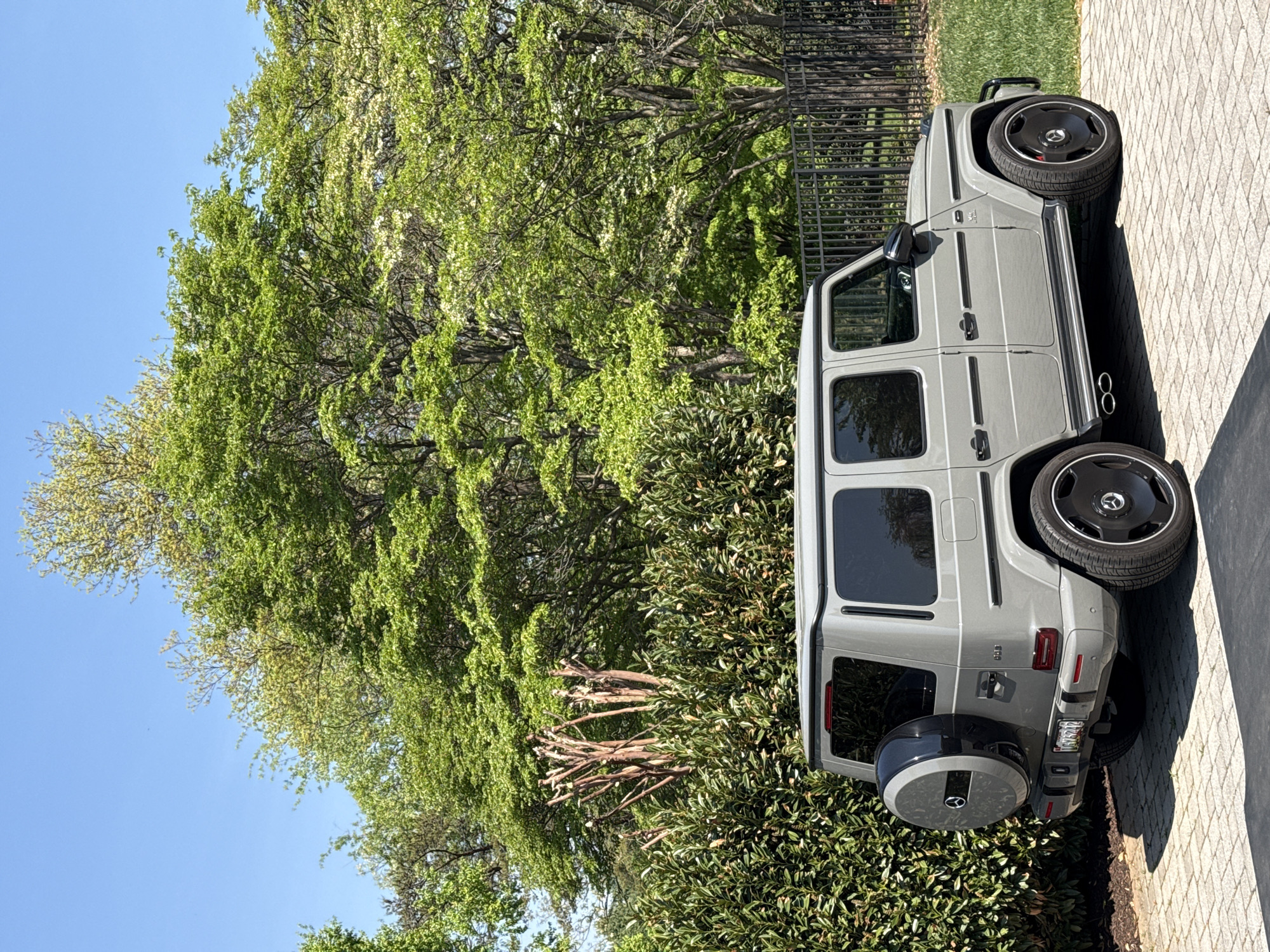 A light gray Mercedes-Benz G-Wagen (G-Class SUV) is prominently featured, parked on a paved driveway made of light-colored paver stones. The vehicle is seen from its passenger side, with the front facing away from the viewer towards the left. It has distinct black multi-spoke wheels with Mercedes-Benz star logos in the center, and its side-exit exhaust pipes are visible beneath the rear passenger door. A "V8 BITURBO" badge is present on the front fender. The rear of the vehicle shows a covered spare tire mounted on the door, also featuring the Mercedes-Benz star logo. A partially visible license plate on the rear appears to be from the United States.
The car is situated outdoors on a clear, sunny day, suggested by the bright light and distinct shadows. The background is dominated by lush green trees with fresh, light-green foliage, indicating springtime or early summer. A vibrant blue sky is visible above the trees. Behind the car, a black metal fence separates the driveway from a grassy area. In front of the rear of the car and extending into the lower frame, there are dense green bushes. The overall setting suggests a well-maintained residential or suburban environment, consistent with the provided location context of Potomac, United States of America. No people are visible in the image, and no specific activity is taking place beyond the car being parked.