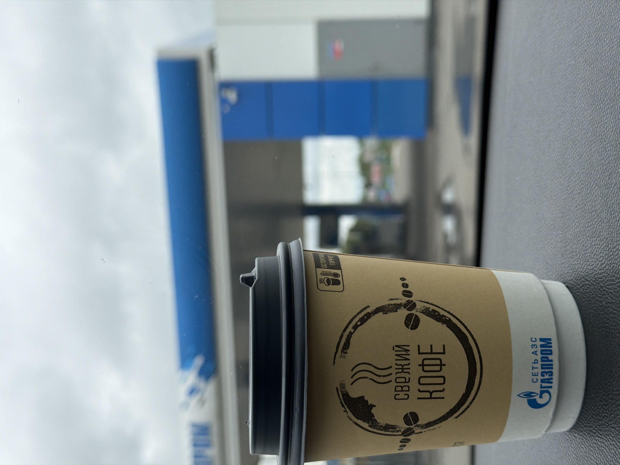 A close-up, eye-level shot reveals a disposable coffee cup positioned on a dark, textured vehicle dashboard, with a blurred Gazprom gas station in the background under a cloudy sky in Совхозный, Russia. The cup, predominantly light brown/kraft paper, features a black plastic lid. Its design includes a distressed circular emblem with the Russian text "СВЕЖИЙ КОФЕ" (Fresh Coffee), flanked by stylized coffee beans and steam rising from a cup graphic. Above this, a small icon with a thermometer and a hot beverage symbol is accompanied by the Russian warning "ОСТОРОЖНО ГОРЯЧО!" (Caution Hot!). The bottom of the cup displays the blue Gazprom logo and the text "СЕТЬ АЗС ГАЗПРОМ" (Gazprom Gas Station Network) on a white band. The background shows the blue and white structures of a gas station canopy with a partial "GAZPROM" logo visible on the left, and a prominent blue pillar on the right. No individuals are present in the image.