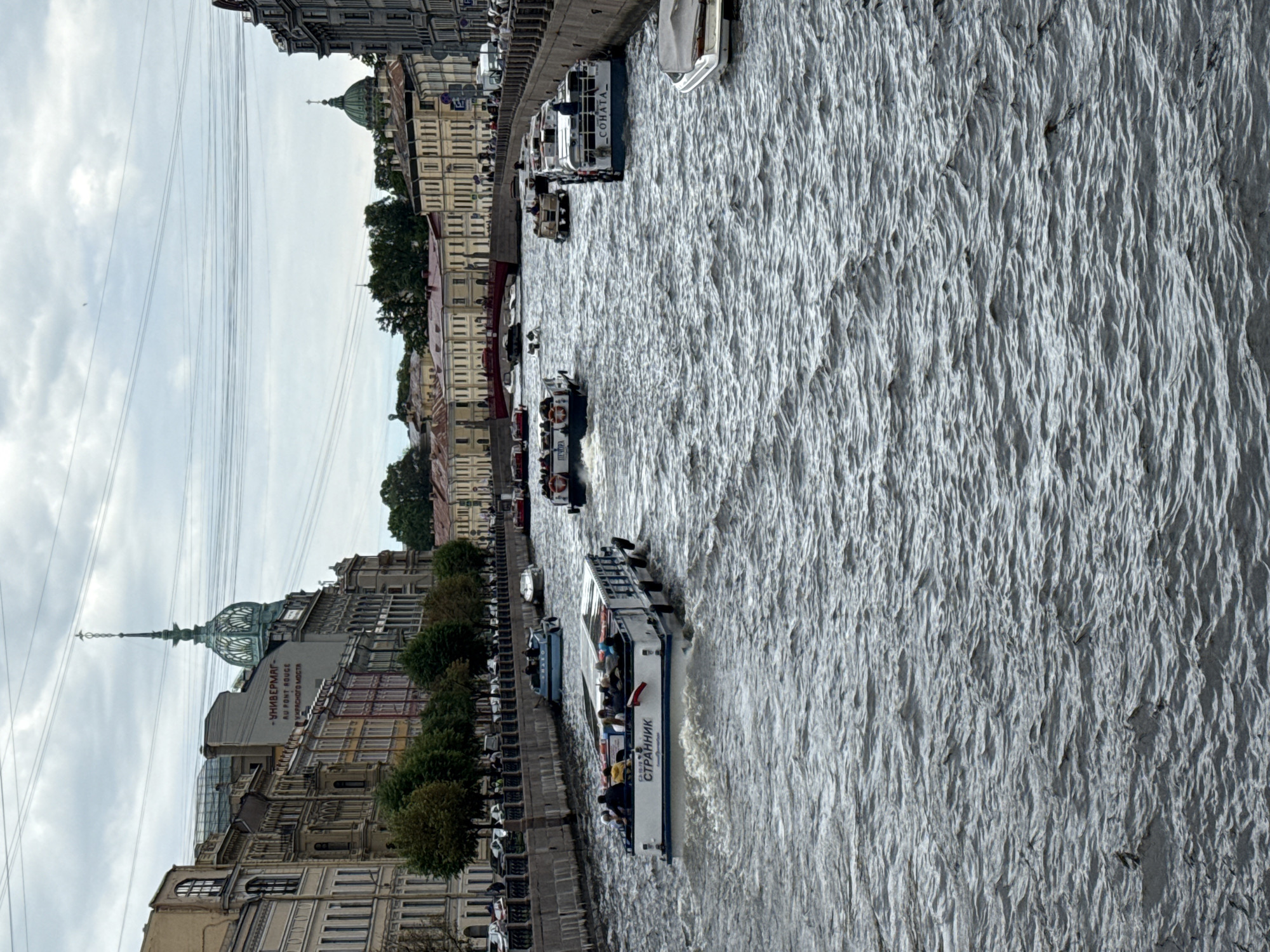 The image captures a canal in Saint Petersburg, Russia, likely the Griboyedov Canal, given the distinctive architecture and the presence of tour boats. The canal is bustling with activity, featuring multiple tour boats navigating the choppy, grey water. One prominent boat in the foreground bears the Cyrillic text "СТРАННИК" (STRANNIK) on its side. Other boats are visible further down the canal, some docked and others in motion.
The canal is flanked by the ornate, multi-story buildings characteristic of Saint Petersburg's historic center. These buildings display a range of architectural styles and colors, with some featuring prominent domes and spires, indicative of churches or administrative buildings. The sky above is overcast with a cloudy, diffused light, casting a muted tone over the scene. Several power lines crisscross the sky, a common sight in urban environments.
People are visible on the tour boats, appearing to be tourists enjoying a canal tour. Some are seated, while others are standing and observing the surroundings. The overall impression is of a vibrant, active urban waterway, a popular tourist attraction in a historic city.