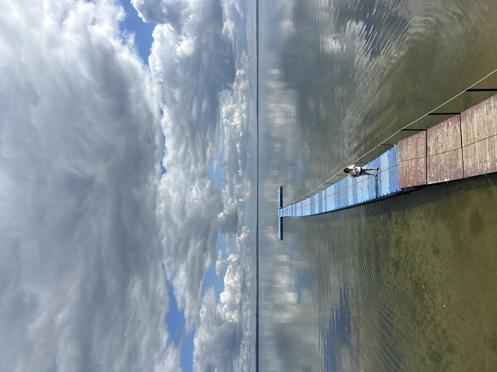 An elevated, vertical, medium shot captures a person standing on a long, narrow pier extending into a calm body of water under a cloudy sky. The pier, constructed of wooden planks and blue floating modular sections, juts out from the sandy shore into the lake. The water is shallow and clear near the shore, gradually becoming deeper and reflecting the overcast sky. The sky is filled with a mix of dark and light gray clouds, interspersed with patches of blue sky. The horizon is a distant, blurred line where the water meets the land. A single person, a woman wearing a light-colored top and shorts, stands on the blue section of the pier, facing away from the camera, looking out at the water. The scene suggests a tranquil, possibly overcast day at a lake or reservoir. The location is identified as Нарацкі сельскі Савет, Belarus.