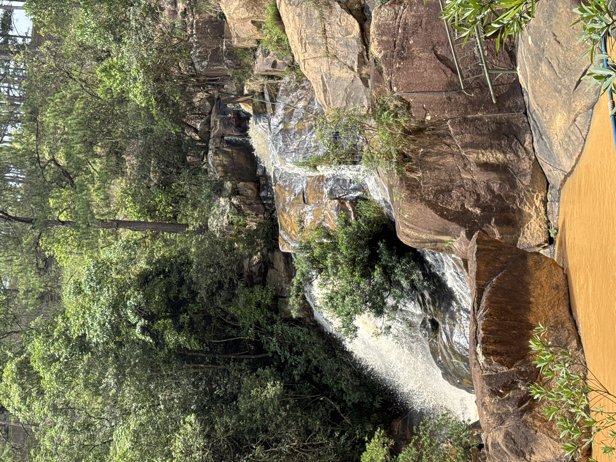 The image shows a waterfall cascading over rocks and boulders. Lush green trees and vegetation surround the waterfall. A ladder can be seen on the right side of the image, attached to the rocks. The location is likely in a natural setting, possibly near Lang Biang - Da Lat, Vietnam, known for its waterfalls. The water appears to be flowing strongly.