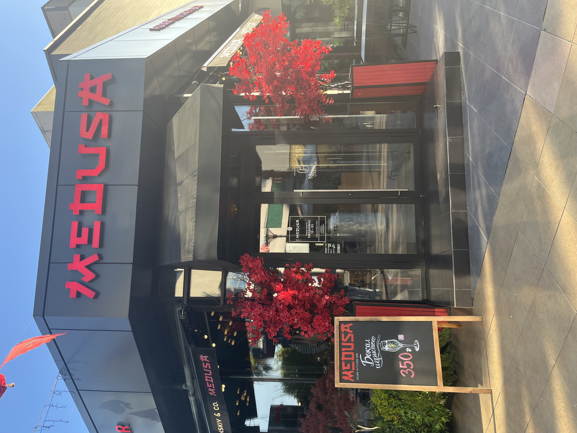 The image shows the exterior of "MEDUSA," an Asian Kitchen Bar located in Sochi, Russia. The restaurant's name is displayed in red capital letters both vertically on the building's façade and horizontally on a chalkboard sign standing in front of the entrance. The entrance has a black awning and glass doors, flanked by red decorative structures resembling trees. A chalkboard sign advertises a glass of sparkling wine for 350 rubles. The building is modern, with a dark gray exterior. The street is paved with light-colored tiles.
