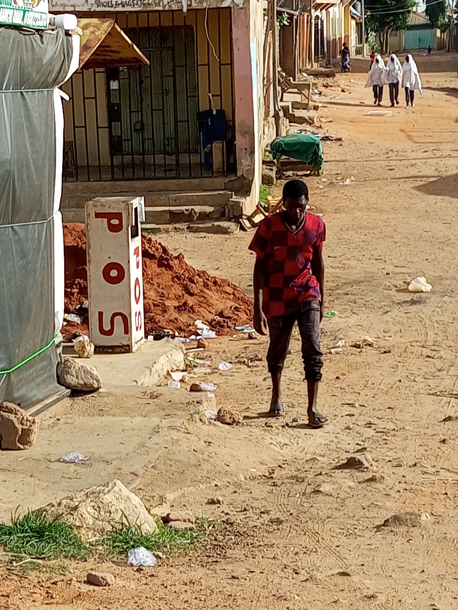 The image depicts a street scene in Gombe, Nigeria. A young Black man in a red and black checkered t-shirt and dark pants is walking towards the right side of the frame on a dirt road. In the foreground, to the left, a large object covered in dark gray material is partially visible. Next to it stands a white rectangular structure with red lettering that spells "P.O.S." A pile of reddish-brown dirt is situated behind the "P.O.S." structure.
In the background, a street lined with buildings stretches into the distance. On the right side of the street, three individuals wearing white hijabs are walking away from the camera. The ground is unpaved and sandy, with scattered debris and some green vegetation growing in patches. The buildings appear to be a mix of shops and residences, some with visible metal security bars on windows and doors. The overall impression is of a dusty, active urban environment.
