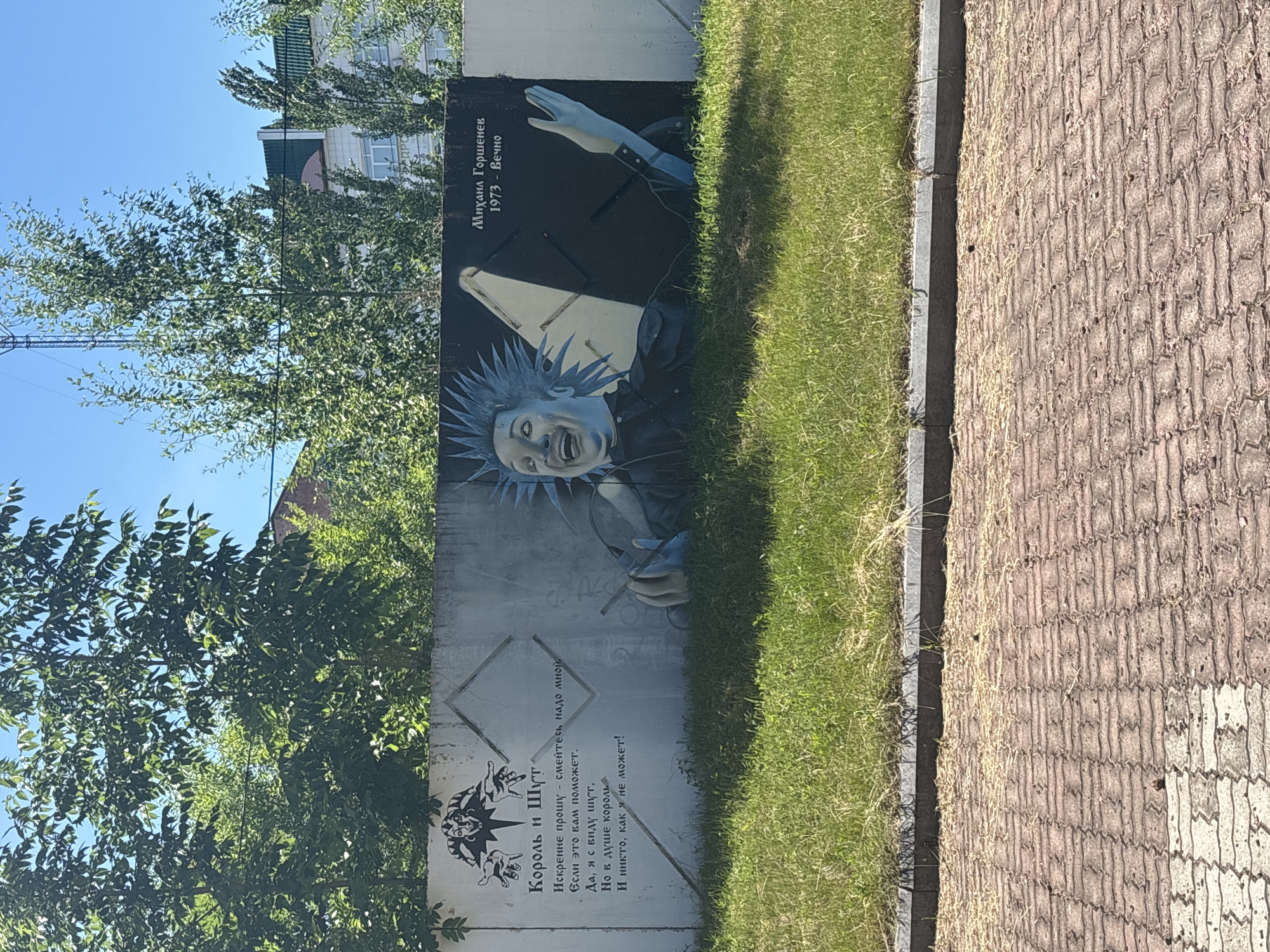 The image shows a mural on a concrete wall in Birobidzhan, Russia, depicting a stylized portrait of a man with spiky blue hair, a wide open mouth, and a cheerful expression. He appears to be holding a cigarette. To the left of the portrait, there is text in Russian, and a smaller graphic of a jester-like figure. The name "Михаил Горшенев" (Mikhail Gorshenev) is written above the portrait, along with the years "1973 - Вечно" (1973 - Forever). The mural is situated outdoors, with green grass and trees in the foreground and background, and buildings visible behind the trees. The wall itself appears to be weathered. The ground in front of the wall is paved with brick-like material. The sky is clear and blue.