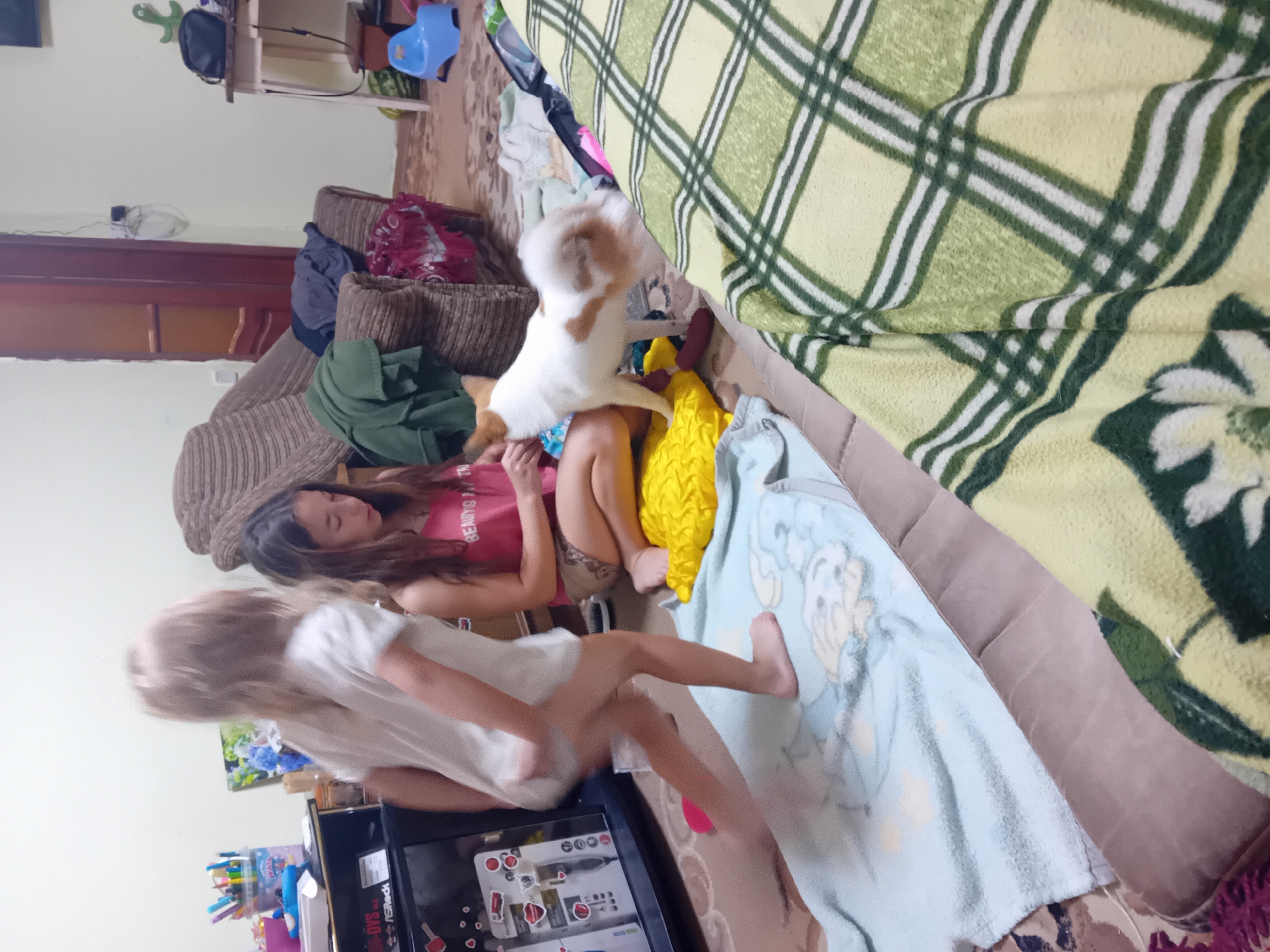 This media file depicts a domestic scene, likely indoors, with two young girls and a small dog. The setting appears to be a living room or a similar communal space, characterized by soft furnishings such as couches and blankets. One girl, with long dark hair, is lying down on a couch, wearing a pink tank top. The second girl, with lighter, shorter hair, is standing and leaning forward, wearing a white t-shirt. A small, fluffy dog, primarily white with brown patches, is perched on the couch near the dark-haired girl, seemingly interacting with the two children.
The girls are dressed casually, suggesting a relaxed, everyday situation. The presence of a dog indicates a home environment where pets are part of the family. A prominent green and yellow plaid blanket covers a portion of the couch, adding to the cozy atmosphere. There are also other blankets and cushions visible, some with patterns, further contributing to the domestic setting. Scattered items like a yellow object, possibly a cushion or toy, and a light blue blanket with a cartoon character motif are also visible. In the background, glimpses of furniture, including a table with items on it, and a doorway can be seen, hinting at a lived-in space.
The overall mood appears to be one of playfulness and relaxation. The children's postures suggest interaction and engagement, while the dog's position indicates it is part of the activity. The lighting suggests daytime. There is no clear indication of the time of day or weather outside. The image is oriented sideways. Text visible on the pink tank top appears to read "BEAUTY IS TRUTH."