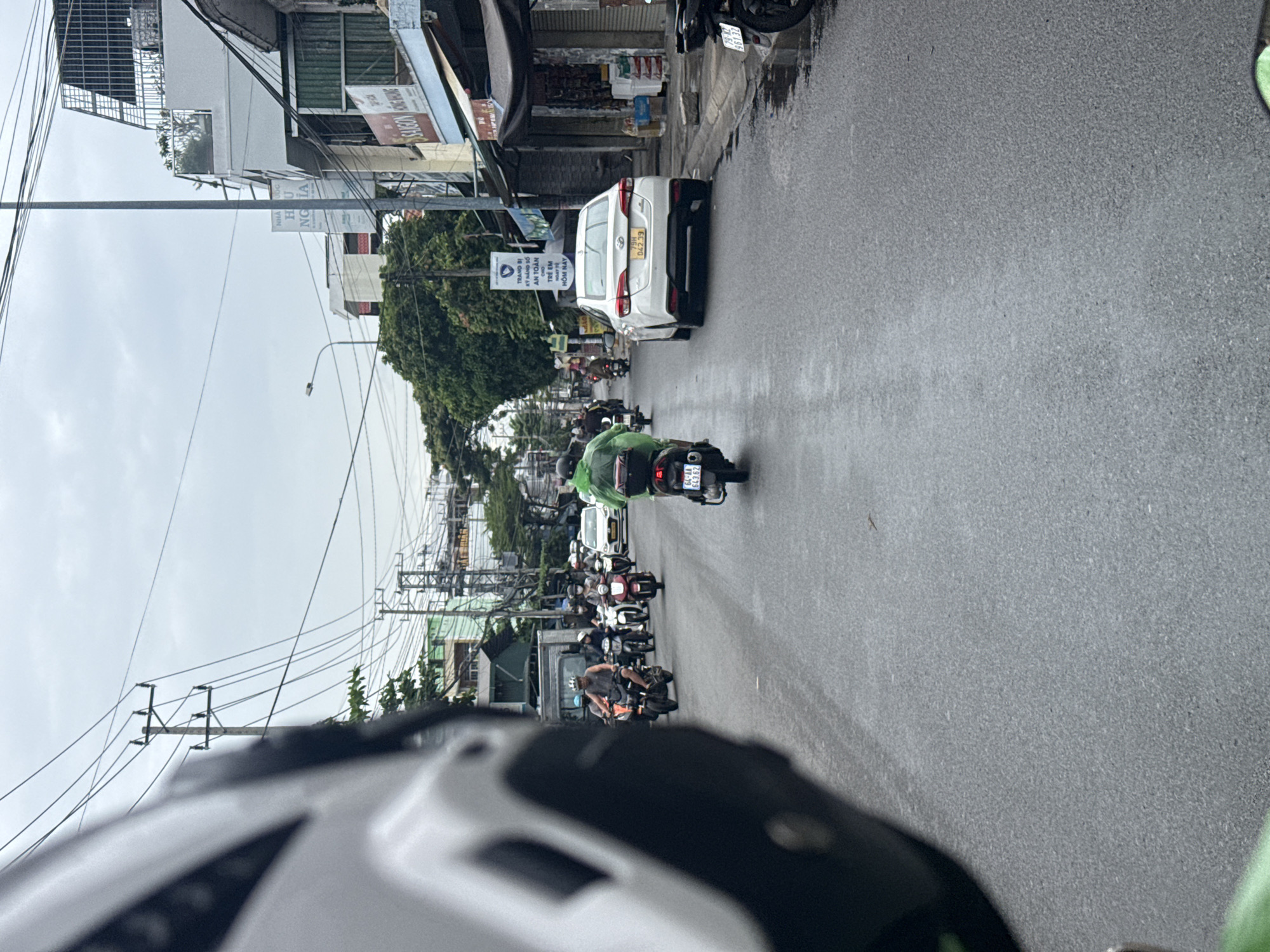 The image shows a street scene in Nha Trang, Vietnam. The street is paved and wet, indicating it might have rained recently or there is standing water. Several motorcycles and one white car are on the road. People are riding the motorcycles, some wearing helmets and casual clothing. The car is a sedan with a license plate that reads "79H 047.39". Buildings line the street on both sides, with shops and some residential structures visible. Power lines and utility poles are prominent overhead, a common sight in Vietnamese cities. The sky is overcast, suggesting a cloudy day. The perspective suggests the photo is taken from another motorcycle or vehicle, with a portion of a helmet visible in the foreground on the left.