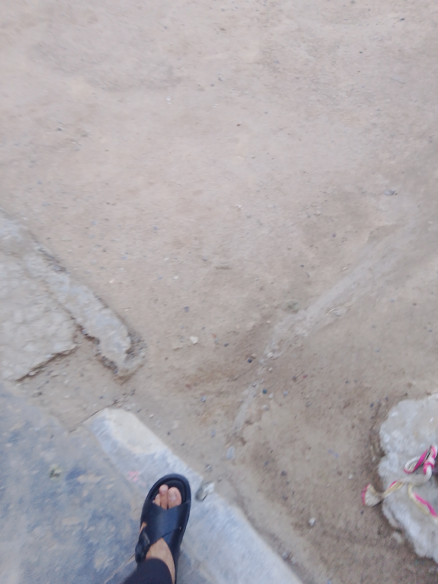 The image shows a close-up view of a person's left foot wearing a black sandal, positioned on the edge of a concrete sidewalk. The ground is a mixture of dirt and small stones, with some patches of lighter colored concrete. To the right of the foot, there is a fluffy white object with pink and yellow rope-like material, possibly a toy or a piece of fabric. The overall environment appears to be an outdoor, possibly urban setting. The photograph's perspective is low, looking down towards the foot and the ground. The specific location in Mansehra, Pakistan cannot be determined from the visual information alone.