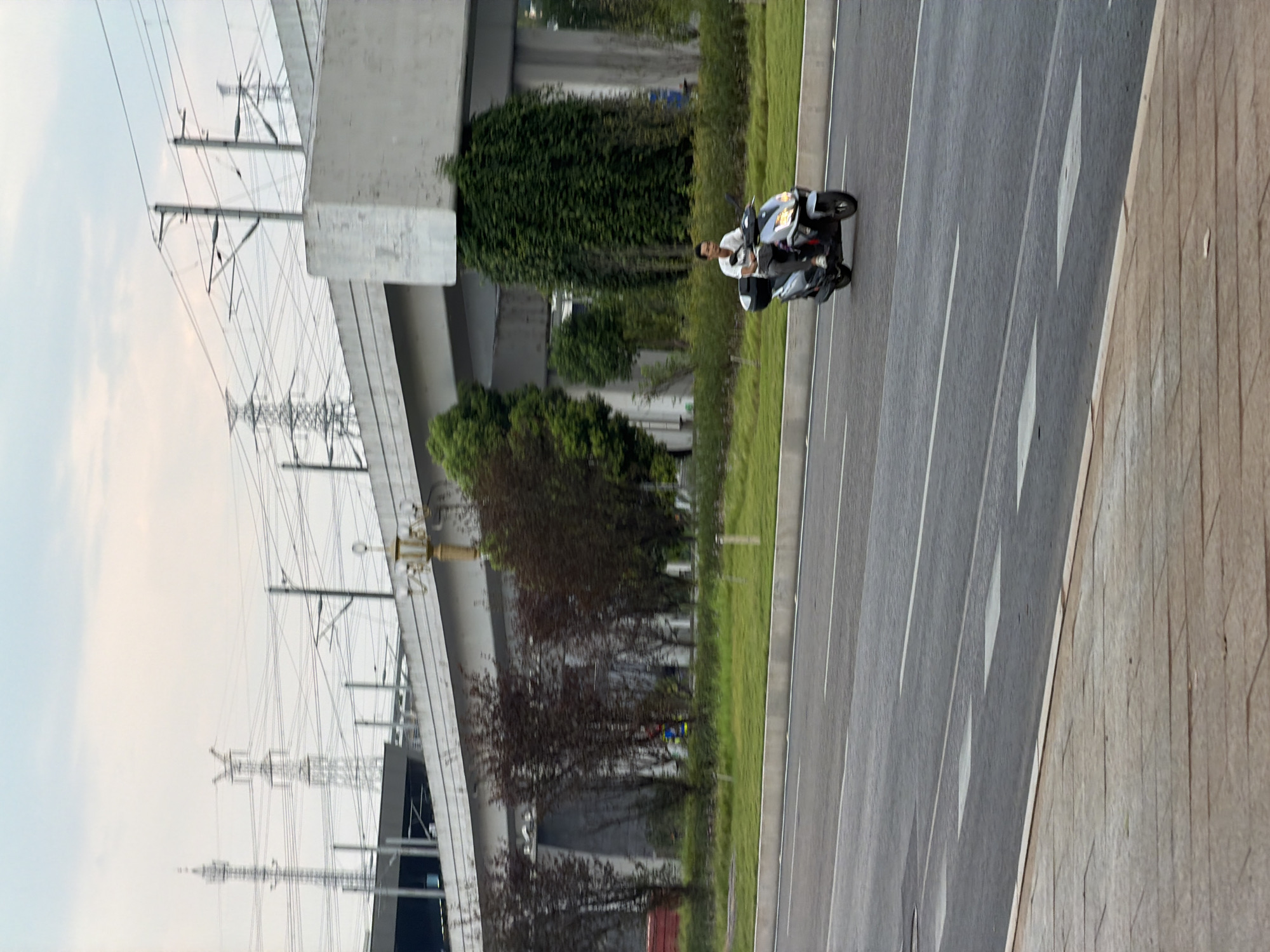 A lone man on a scooter rides along a paved road in Jiangjin, China. The man, wearing a white short-sleeved shirt and dark pants, is seated on a dark-colored scooter with a white storage box behind him. He is looking straight ahead as he drives. The road is lined with a green grassy median strip. In the background, a concrete overpass stretches across the frame, supported by large pillars. Power lines and tall transmission towers are visible in the distance against a cloudy sky. The scene appears to be an urban or suburban setting with infrastructure for transportation and utilities.